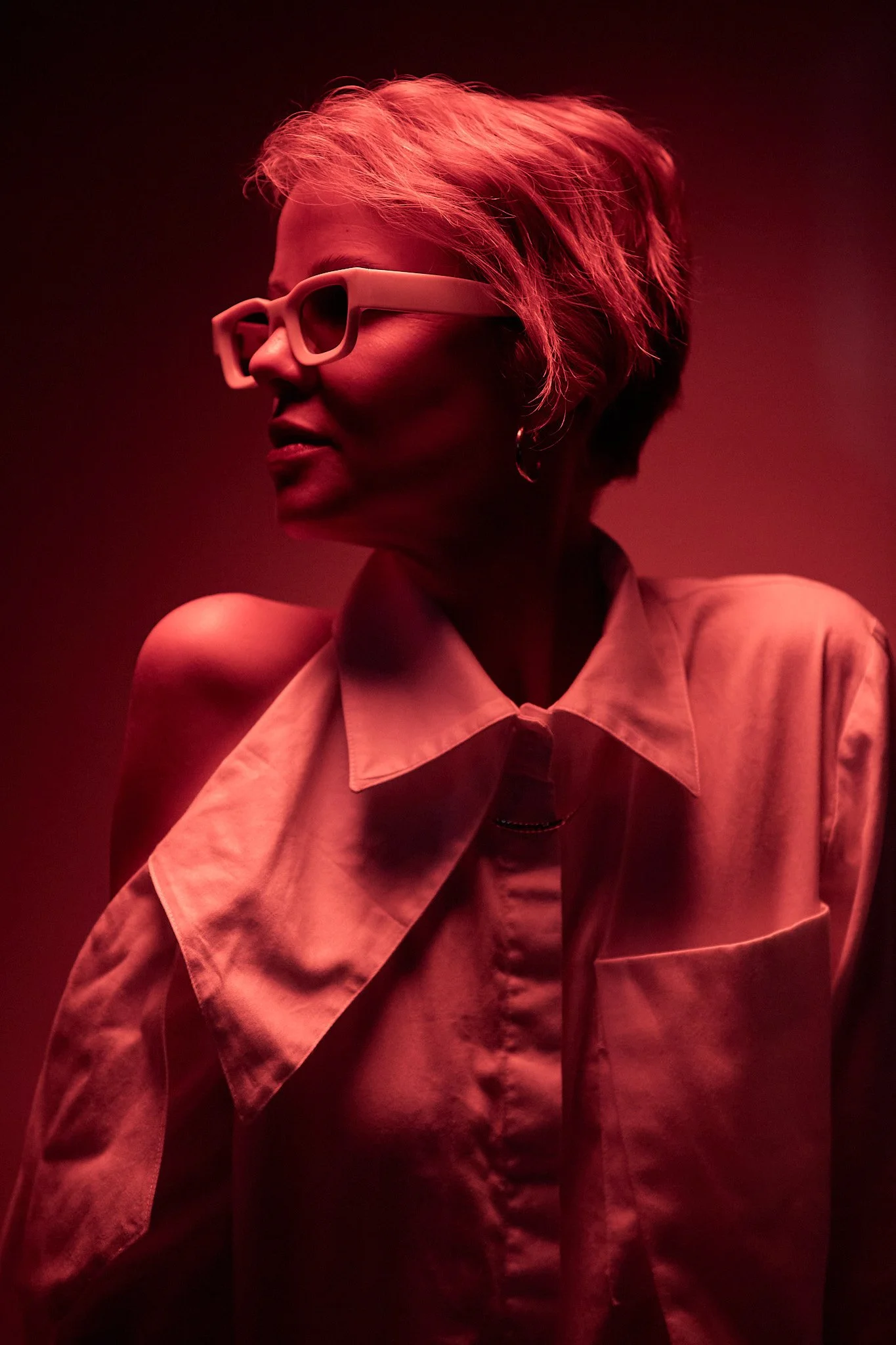 A woman with short, light-colored hair wearing large, white sunglasses and a beige trench coat, illuminated by red lighting, looking to her right against a dark background.