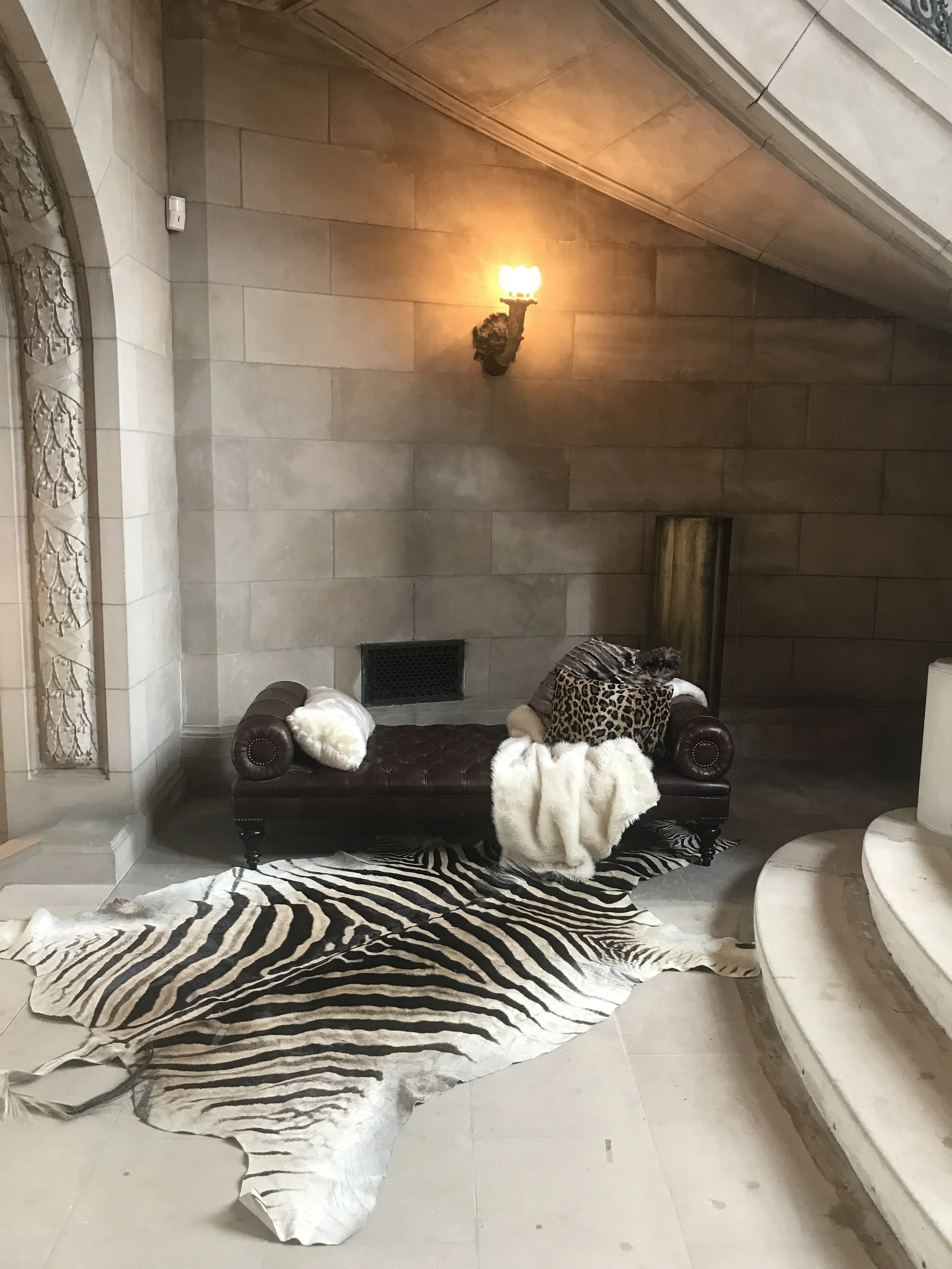 A black leather tufted bench with decorative pillows, including animal print and fur, positioned in a room with stone walls and a zebra print rug. A large zebra-print paper cutout lies on the floor, and a wall-mounted candle-style lamp is lit on the 