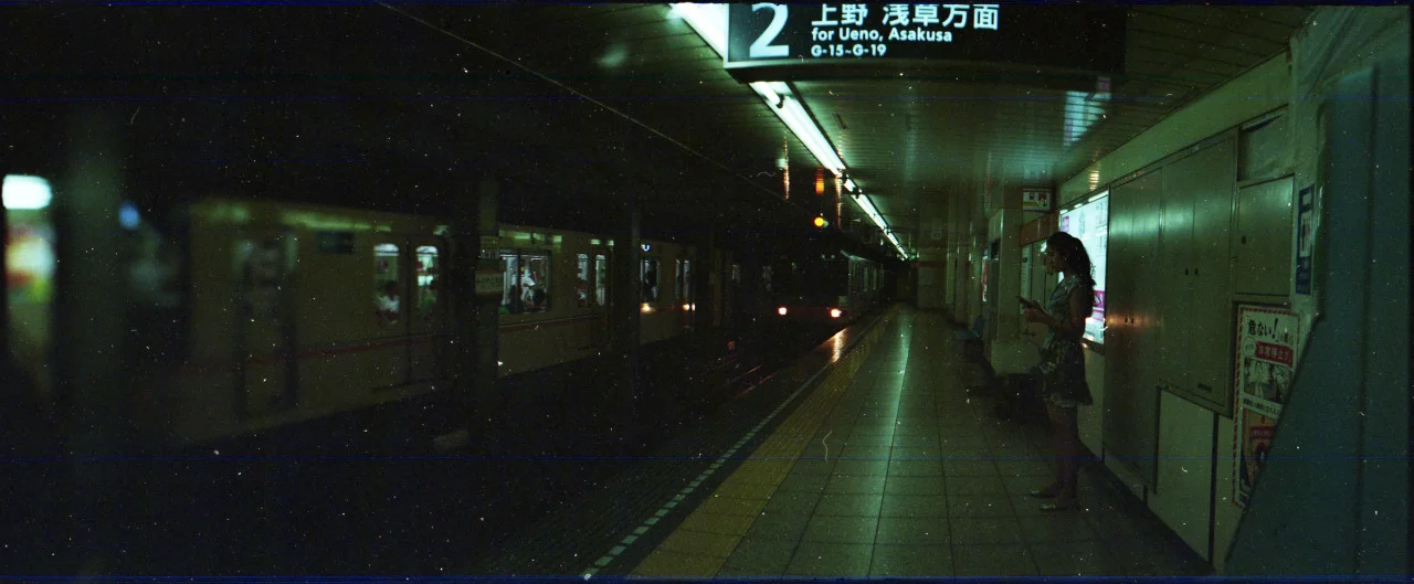 Woman standing on subway platform using her phone, train arriving in the distance, sign indicating platform 2 for Ueno and Asakusa.