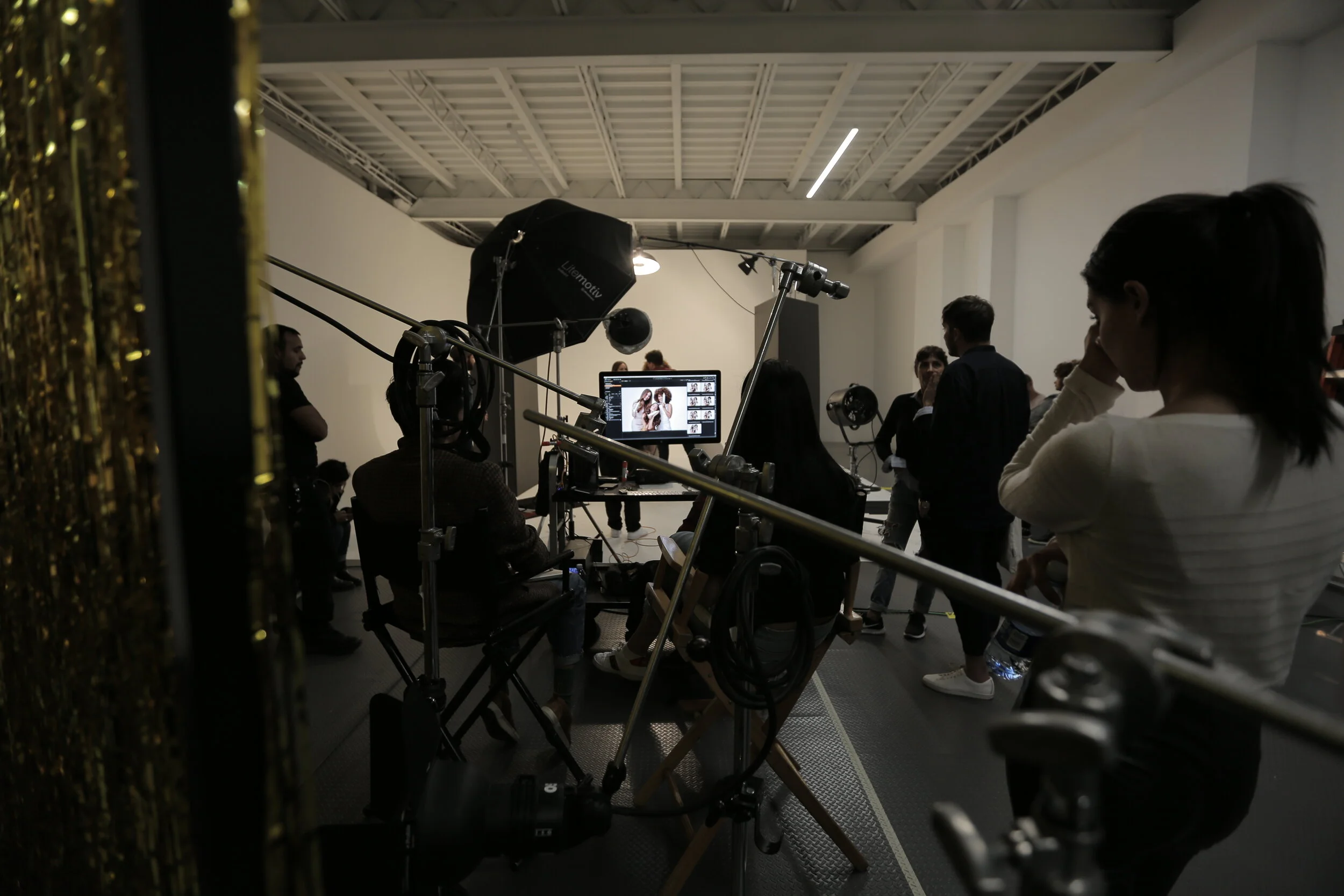 People gathered in a photography studio with a white wall, professional lighting, and a computer monitor displaying photos of two women. Some individuals are engaged in conversation or observing.