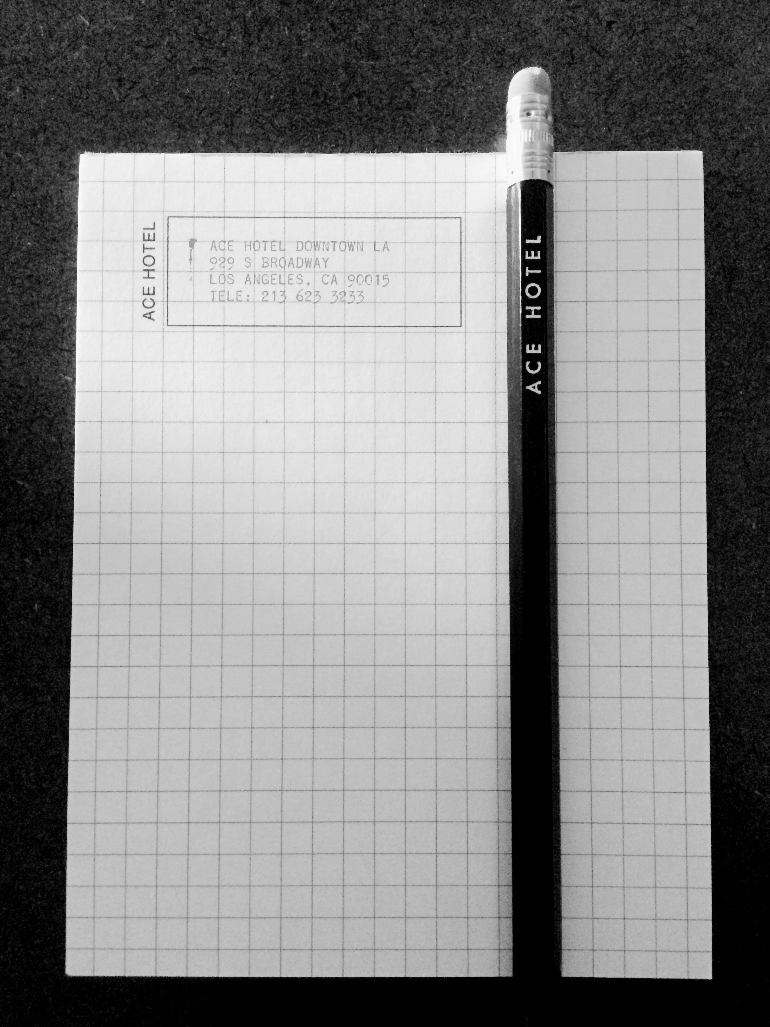 A piece of graph paper with printed hotel address, with a black marker placed vertically on the right side of the paper. The address reads, 'ACE HOTEL DOWNTOWN LA, 929 S BROADWAY, LOS ANGELES, CA 90015, TELE: 213 625 3233'. The paper is on a dark tex