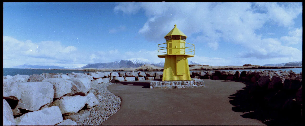 Yellow lighthouse on rocky shoreline with cloudy sky and distant mountains in background.