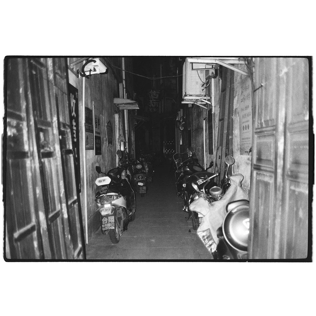 Nighttime alley with parked motorcycles along the sides, wooden walls, and signs, with faint city lights in the background.