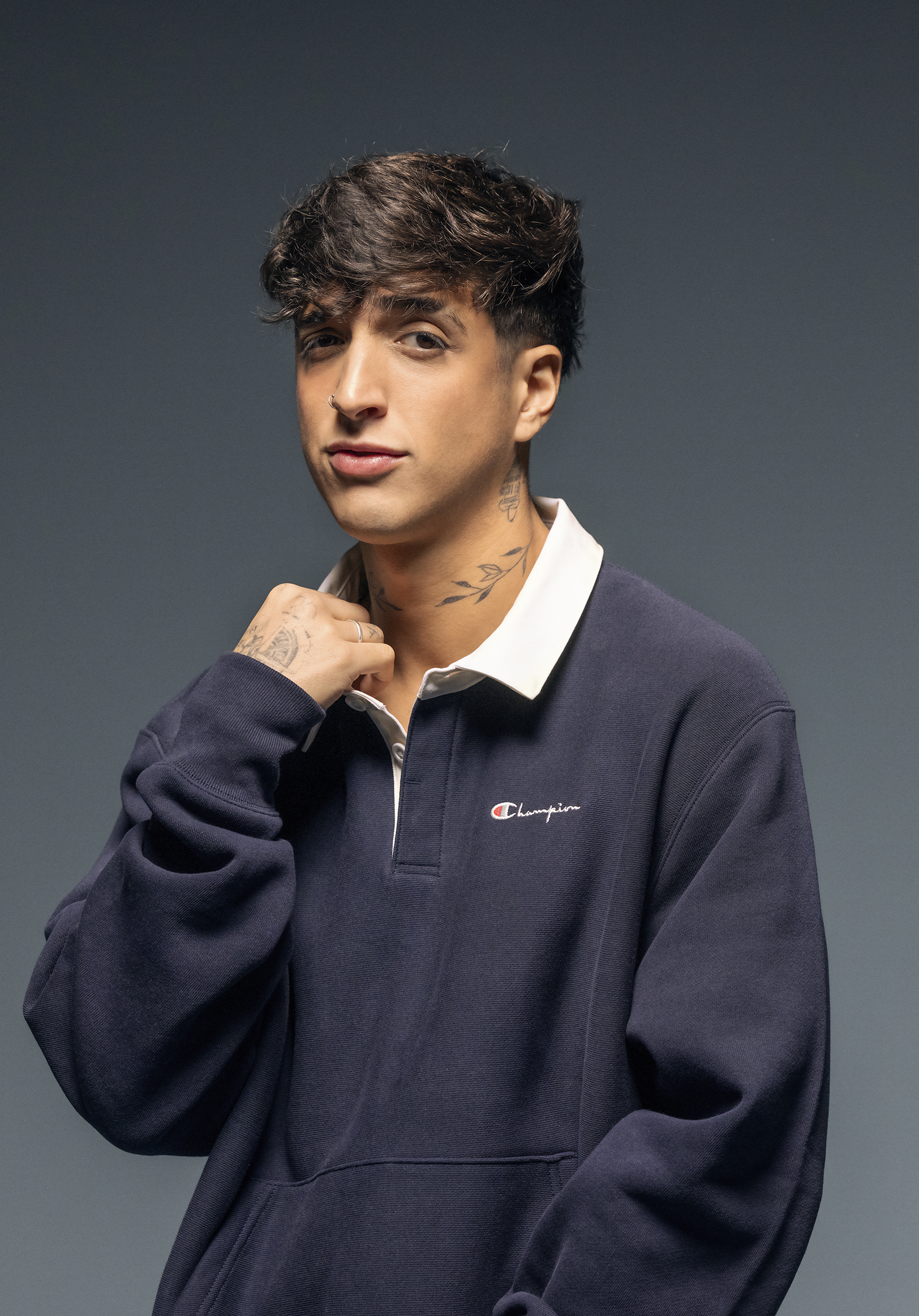 A young man with dark hair, tattoos on his neck and hand, wearing a navy blue Champion sweatshirt and a white collared shirt, posing with one hand near his chin against a plain gray background.