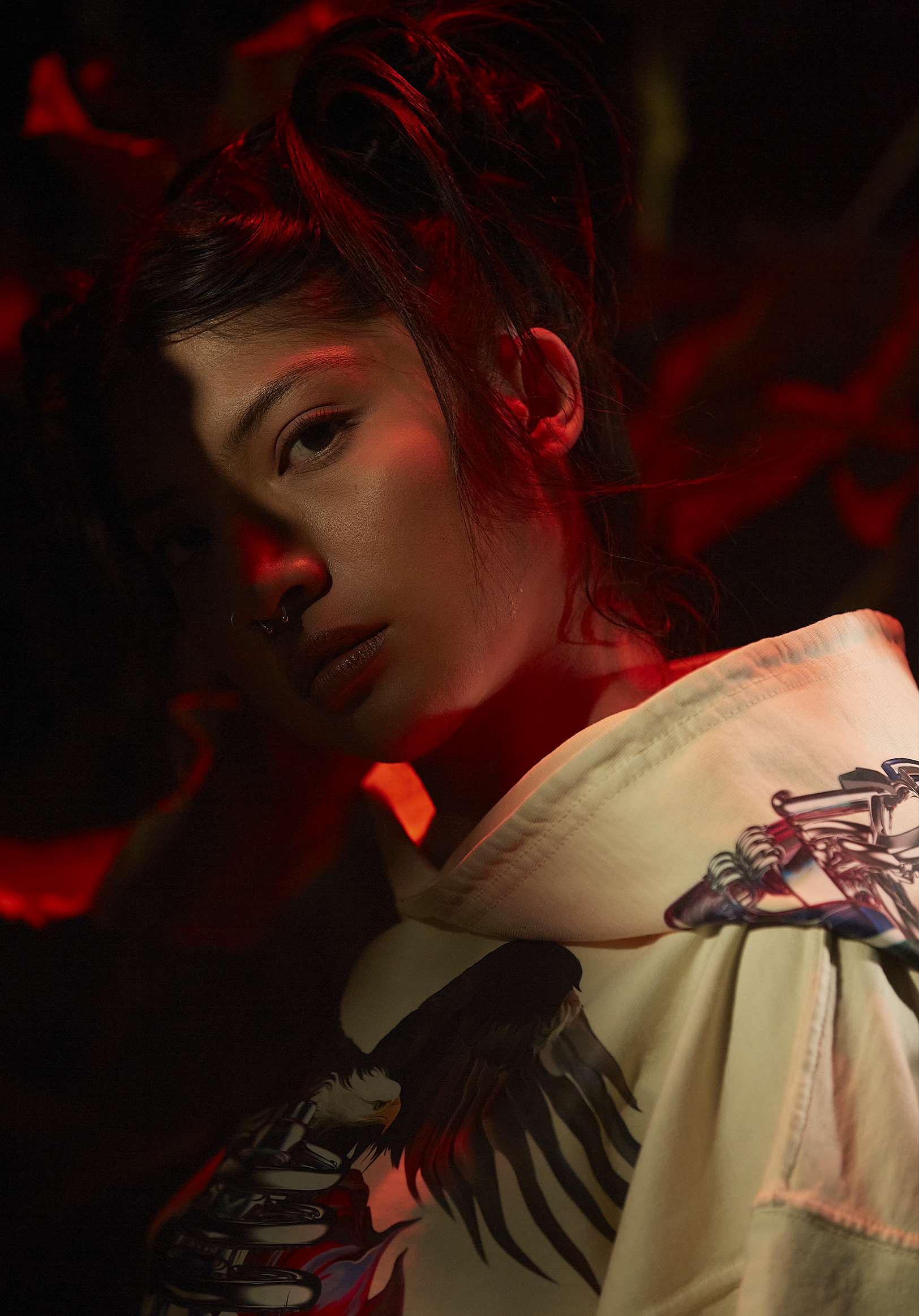 A woman with dark hair and olive skin, wearing a stud nose ring and white hoodie with a colorful graphic, is partially visible in a dark setting with red lighting.
