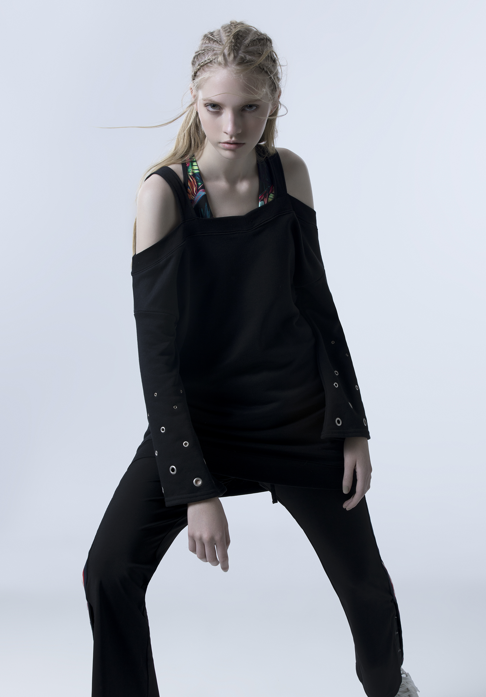 Young woman with blonde hair styled in braids, wearing a black cold-shoulder top with colorful straps, black pants with side details, standing in a pose against a plain light background.