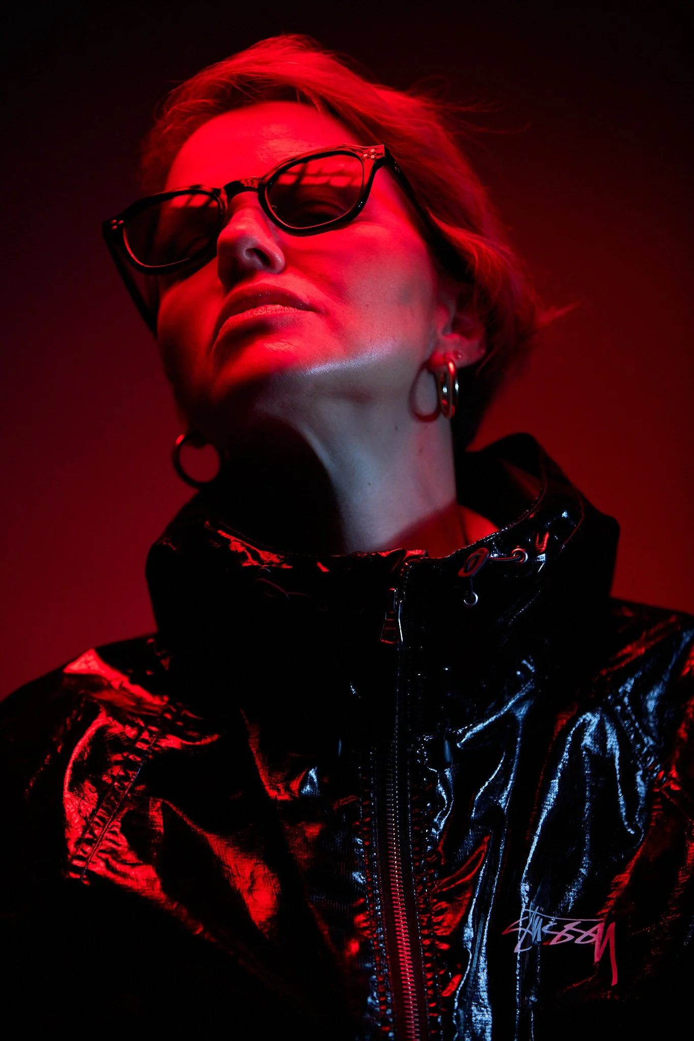 A woman with short, light-colored hair wearing sunglasses and earrings, dressed in a shiny, black, reflective jacket with a high collar, illuminated by red and black lighting.