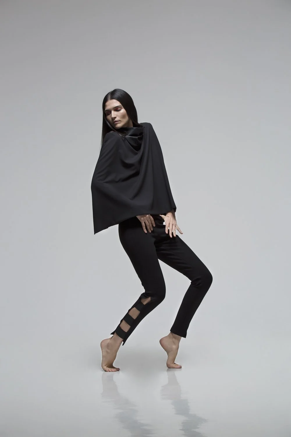 A woman with long dark hair wearing black clothing and standing barefoot against a plain light gray background.