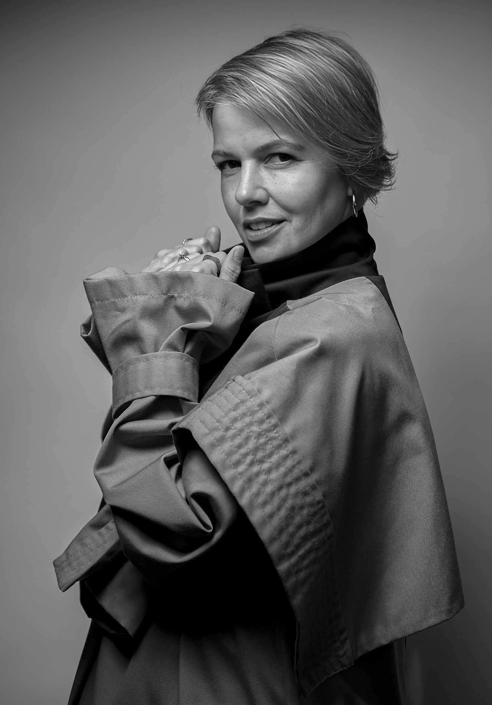 Black and white portrait of a woman with short hair, wearing a turtleneck and an oversized coat, smiling softly and holding her hands near her face.