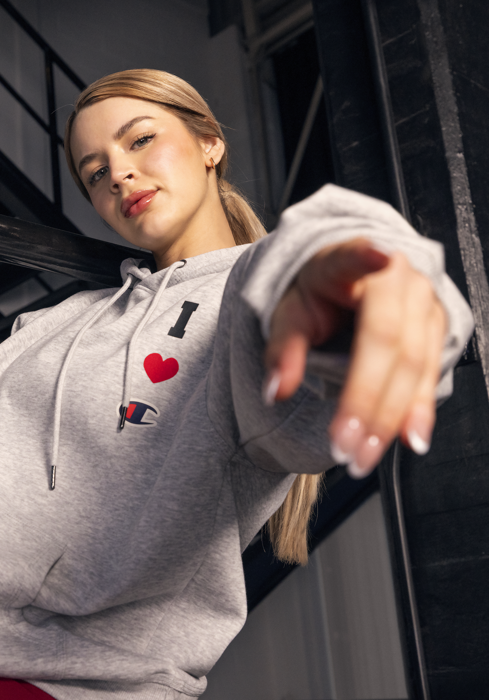 Close-up of a young woman with blonde hair in a ponytail, wearing a light gray Champion hoodie with the text 'I ❤️' on it, smiling and looking at the camera while reaching out with her hand.