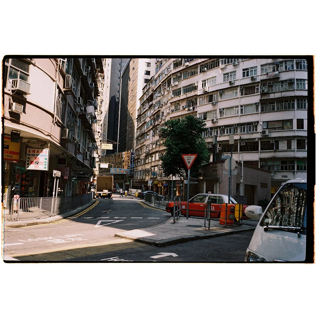 Urban street scene with tall residential buildings, parked cars, a traffic sign, and a tree in the middle of the sidewalk, in a city with high-rise buildings in the background.