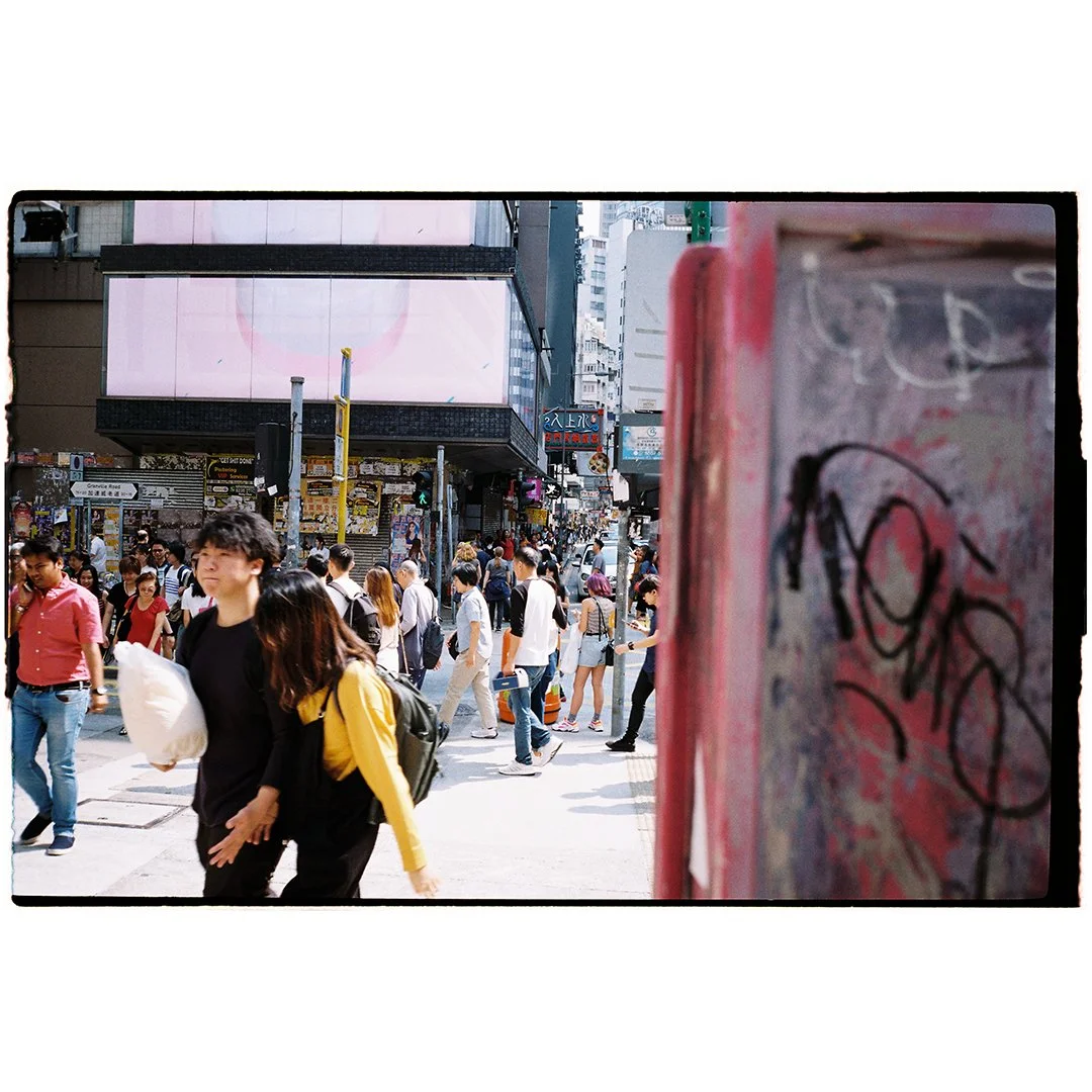 Crowded city street in daylight with people walking, some carrying bags, and a red graffiti-covered object in the foreground.