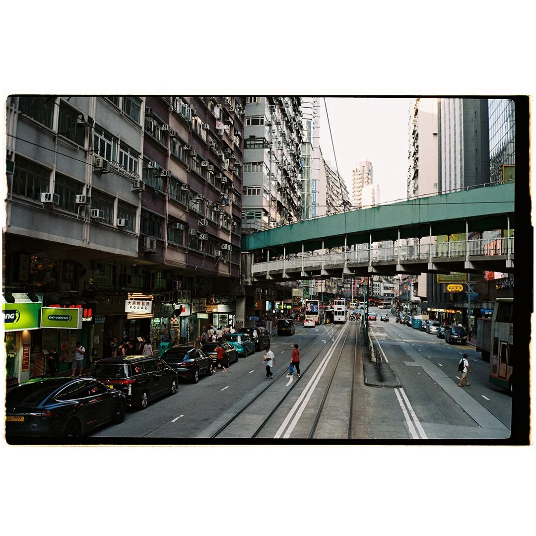 Urban street scene with tall buildings, cars parked along the curb, pedestrians crossing the street, and an elevated green pedestrian bridge.