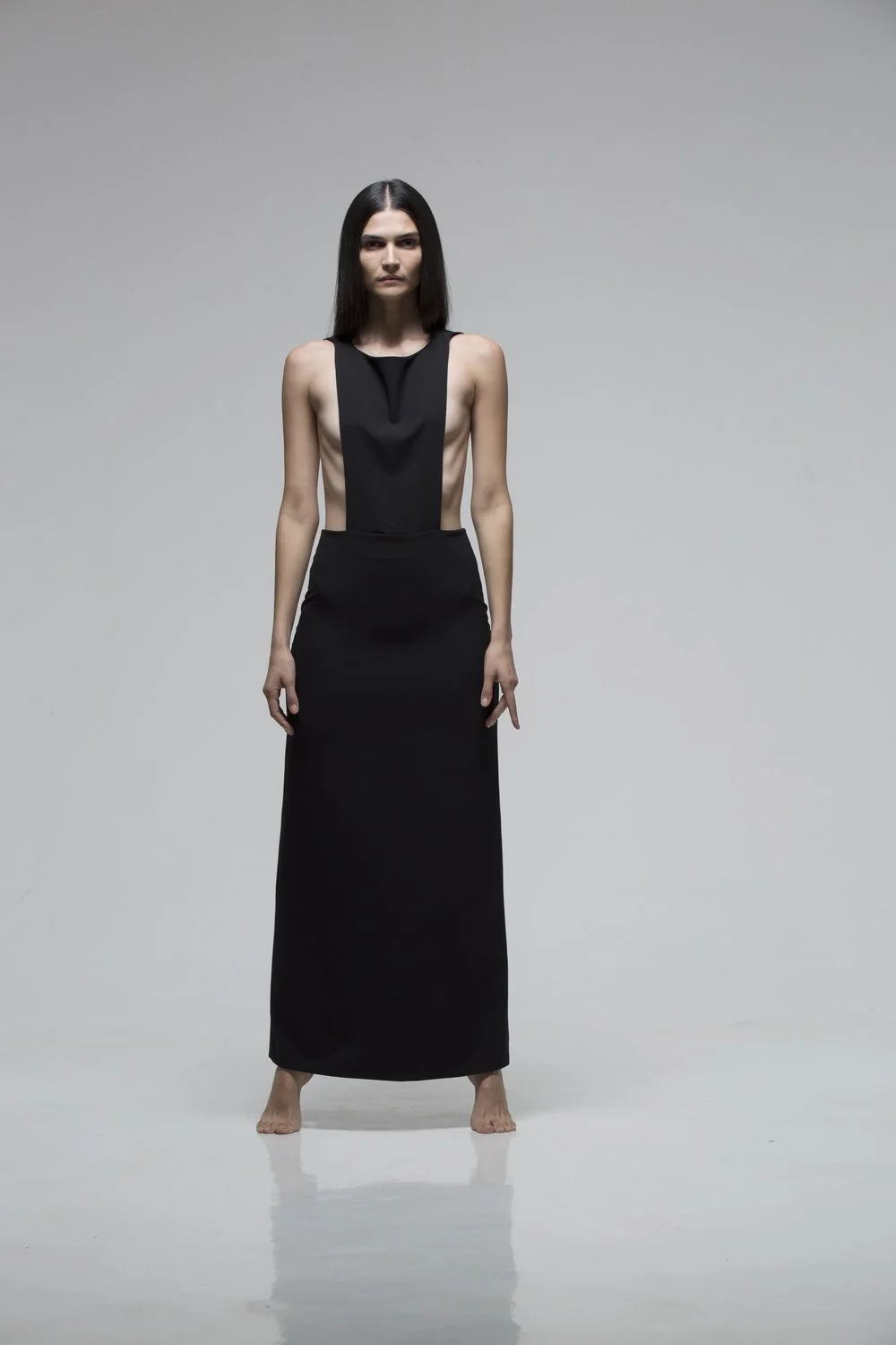 A woman with long dark hair wearing a floor-length black dress with cutouts on the sides, standing barefoot on a plain, light gray background.