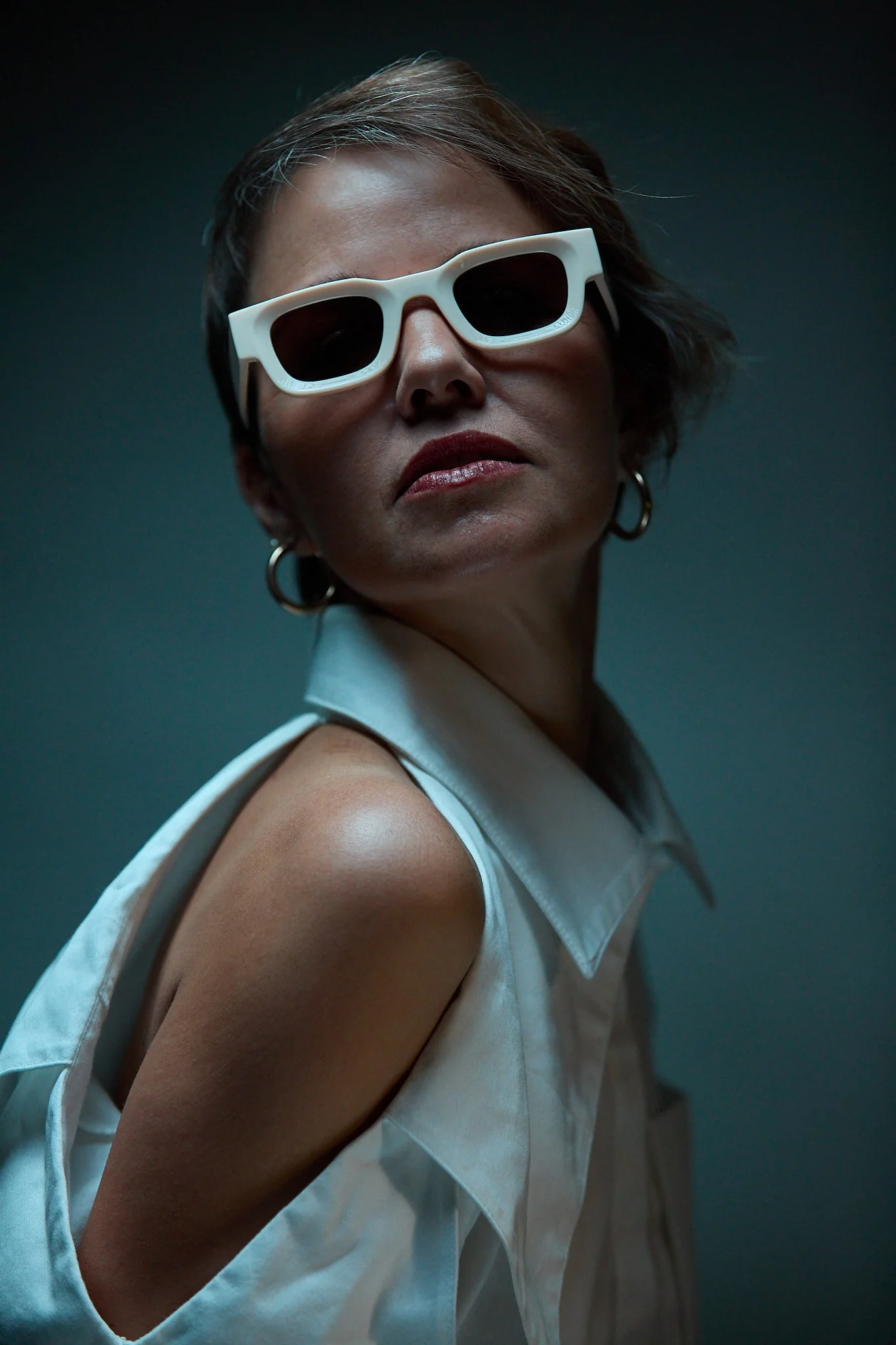 A woman with wavy brown hair wearing white sunglasses with black lenses, a white sleeveless collared shirt, and hoop earrings, posing against a dark background.