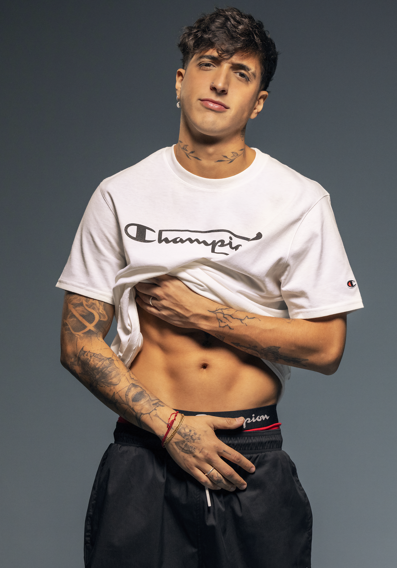 Young man with tattoos smiling slightly, lifting his white 'Champion' T-shirt to reveal part of his torso, wearing black athletic shorts, standing against a gray background.
