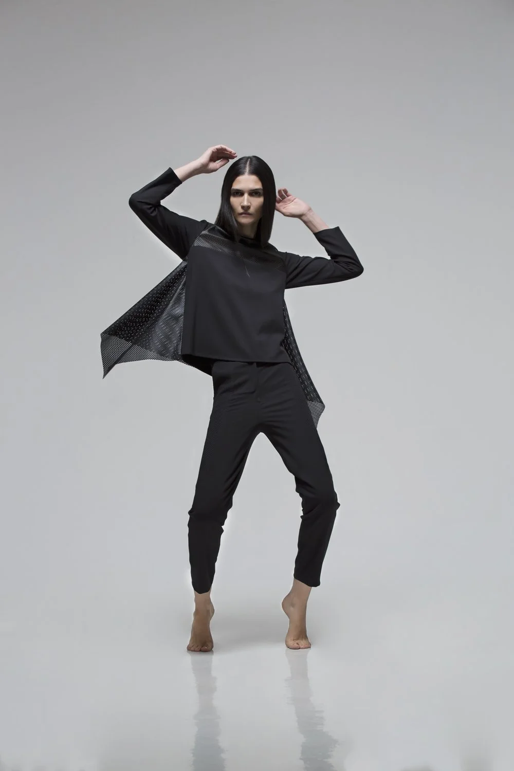 A woman with long dark hair wearing a black outfit with a perforated design, posing barefoot against a plain gray background.