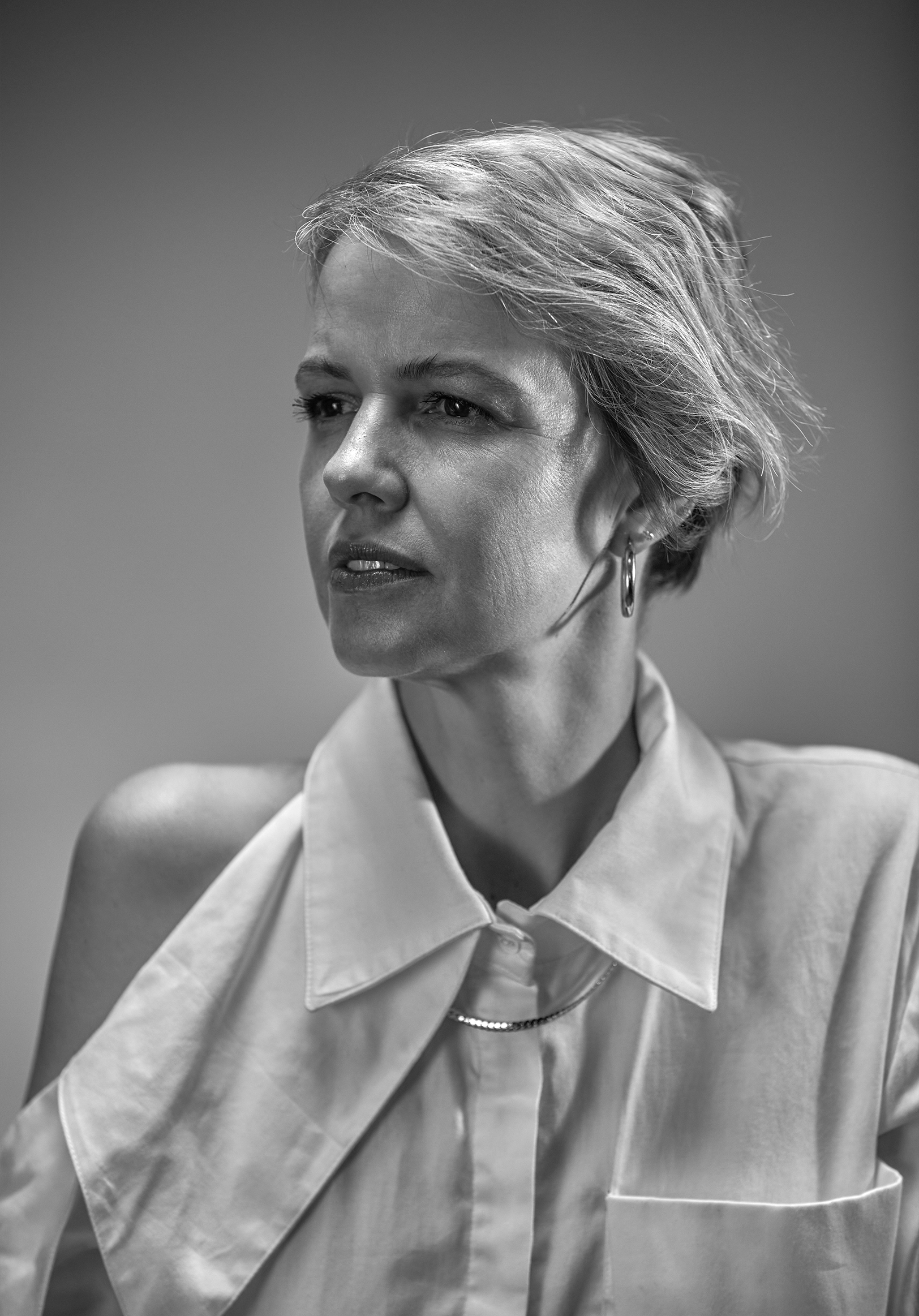 A black and white close-up portrait of a woman with short, tousled hair, wearing a collared shirt, hoop earrings, and a chain necklace, looking to the left.