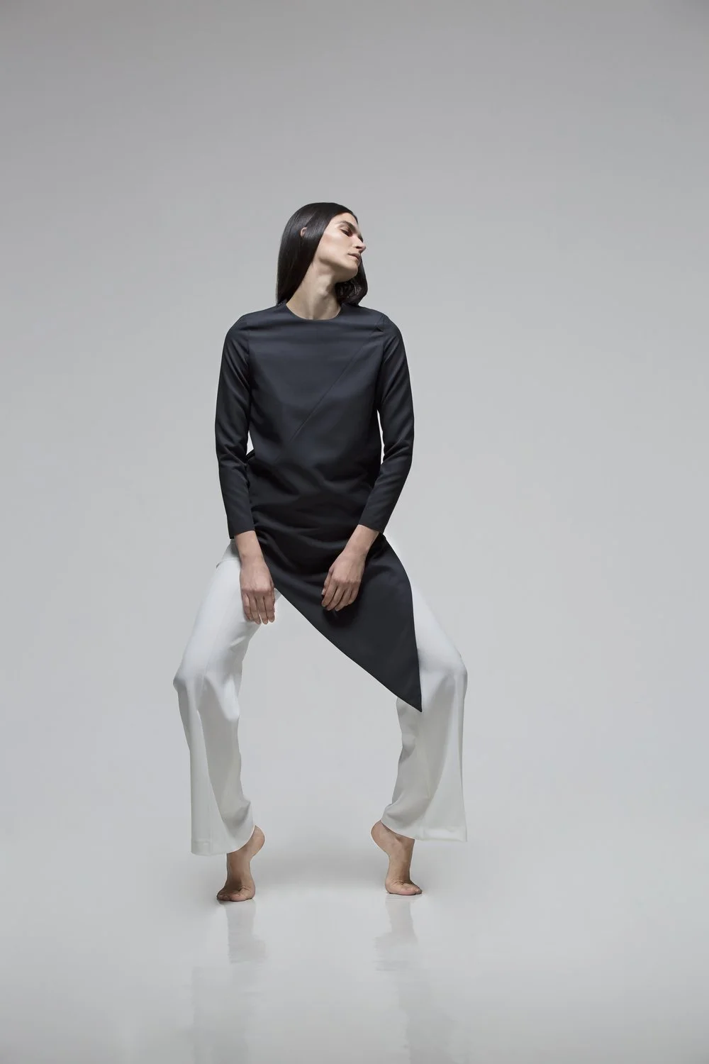 A woman with long dark hair wearing a black top and white pants posing barefoot against a plain gray background.