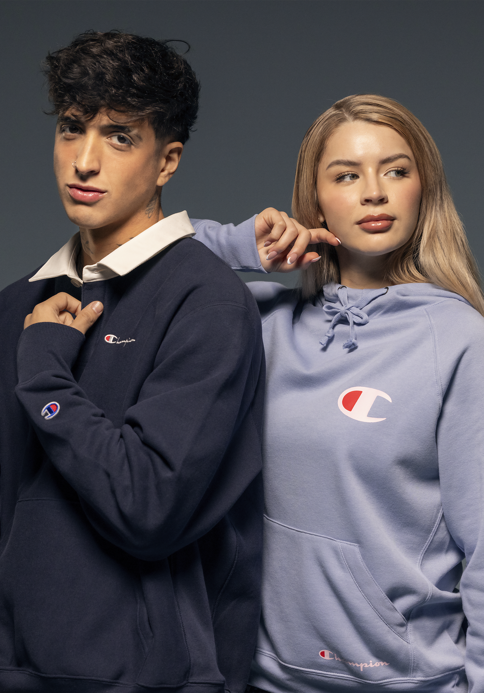 A young man and woman posing together against a gray background wearing Champion hoodies.