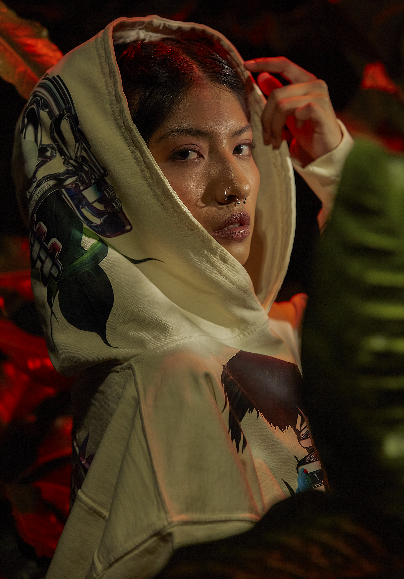 Close-up of a woman with dark hair and septum piercing, wearing a hooded sweatshirt with graphic print, in a dimly lit setting with red and green lighting.