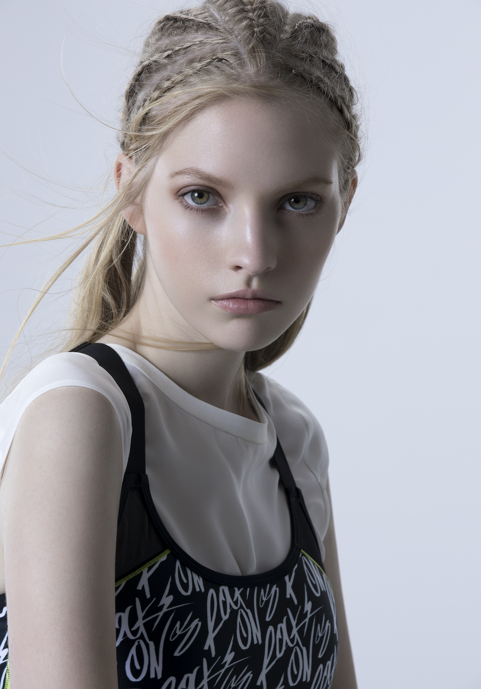 A young woman with blonde hair styled in braids and loose strands, wearing a sleeveless top with graphic text, against a plain light gray background.