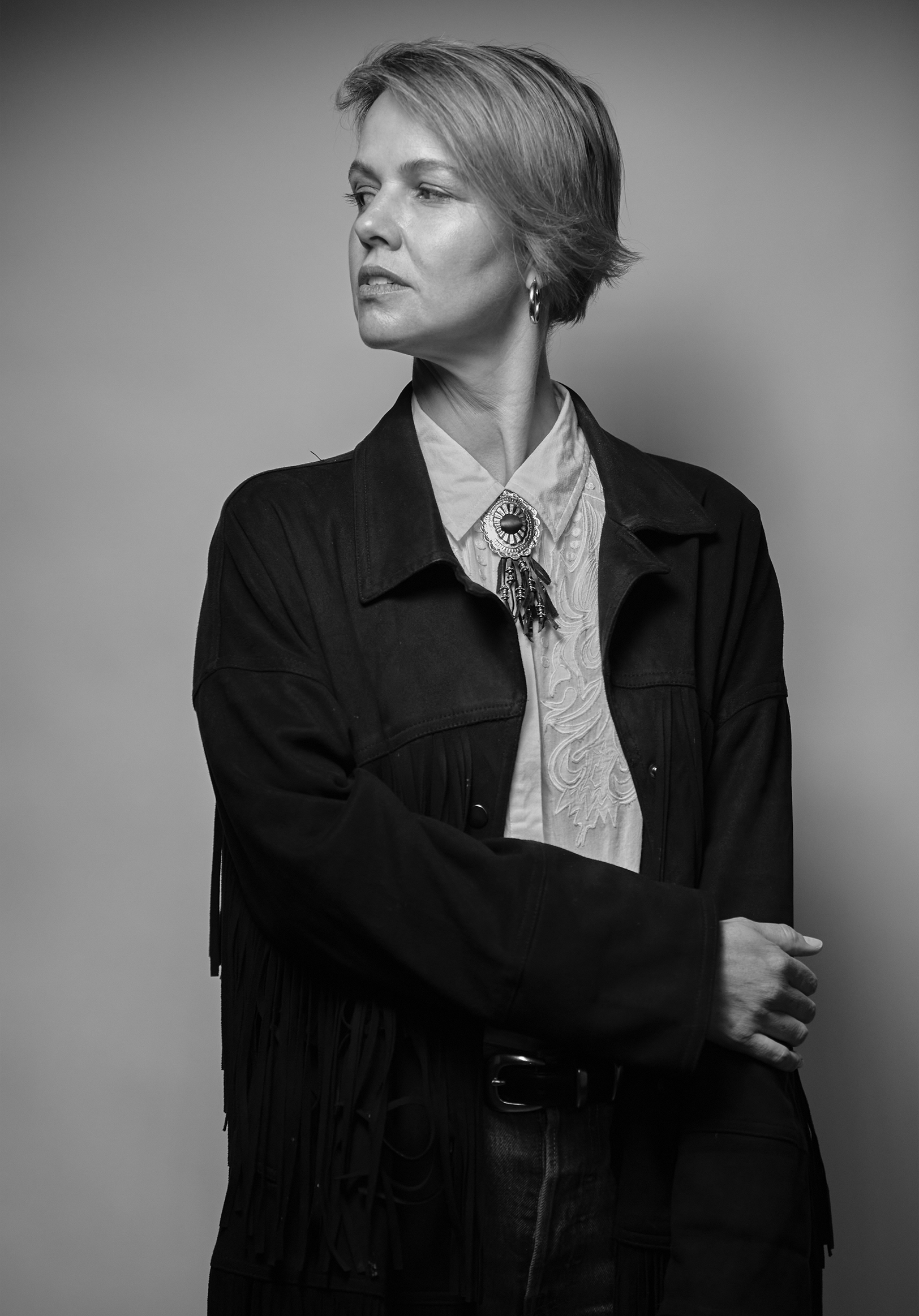 Black-and-white portrait of a woman with short hair, wearing a dark jacket with fringe details, a patterned shirt, and a large decorative brooch. She is looking to her left with her arms crossed.