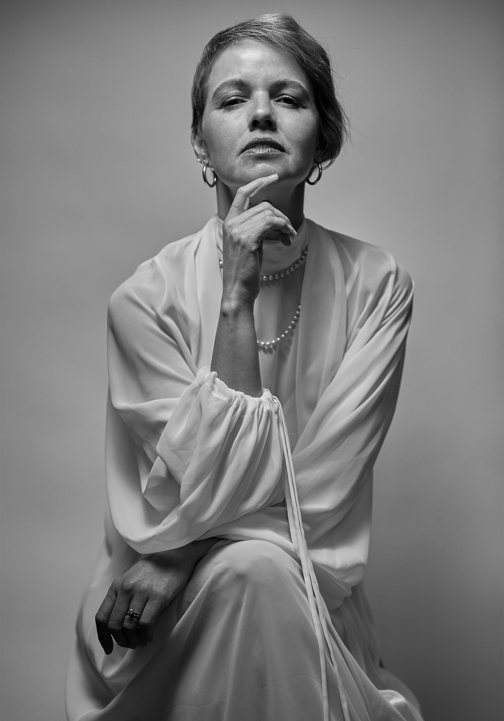 Black and white portrait of a woman with short hair, looking contemplative, wearing earrings, necklaces, and a satin blouse, with her hand touching her chin.