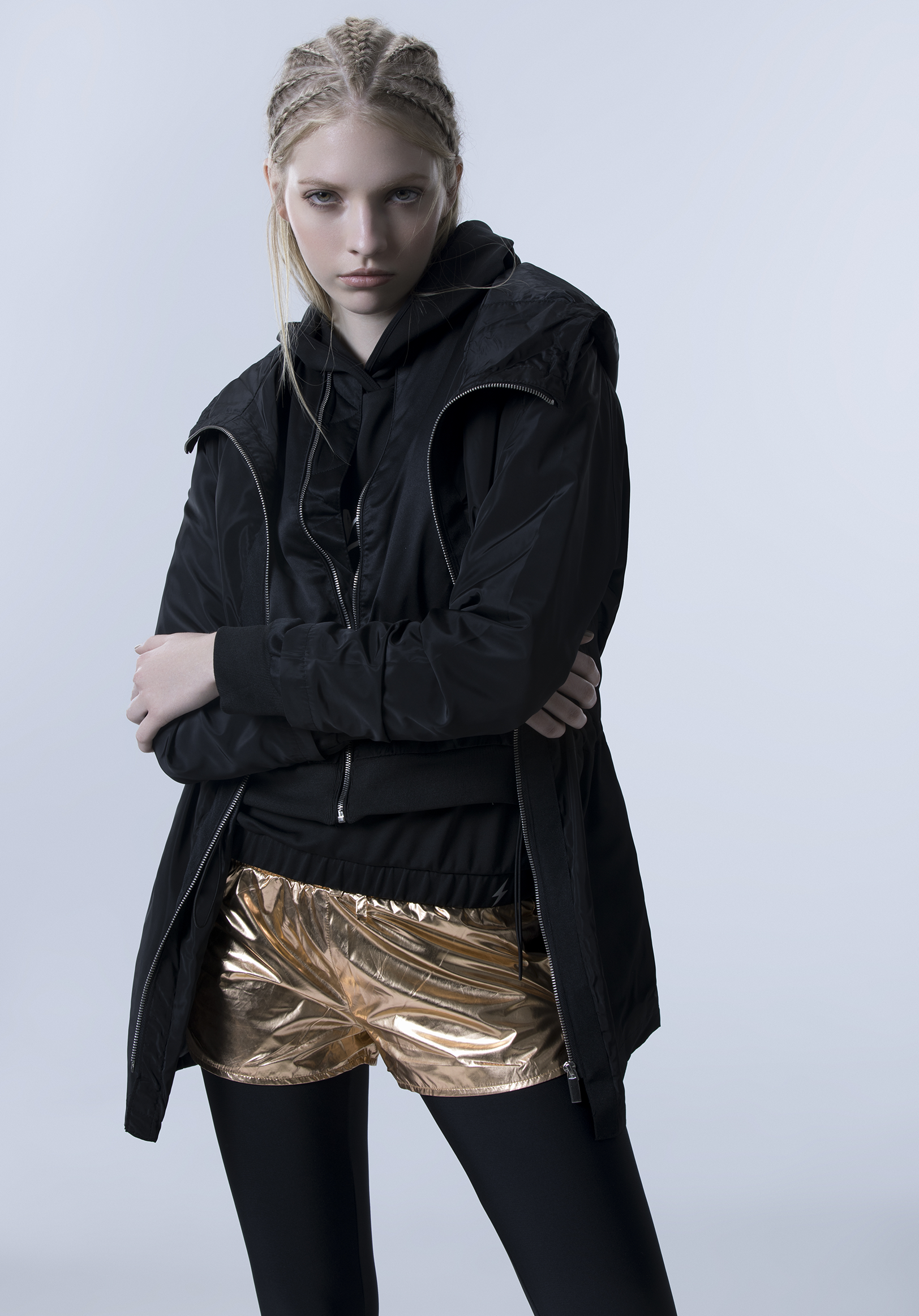 Young woman in a black jacket and metallic shorts with black leggings posing against a plain background.