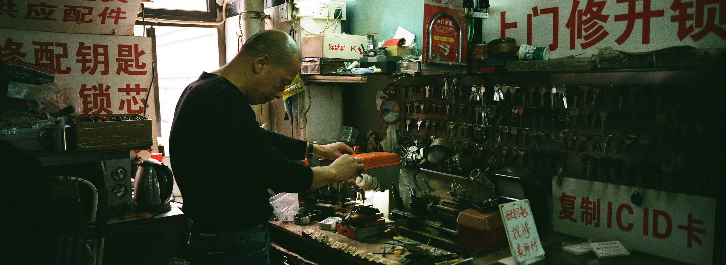 A man working in a locksmith shop surrounded by keys, tools, and security devices, with Chinese signs on the walls.