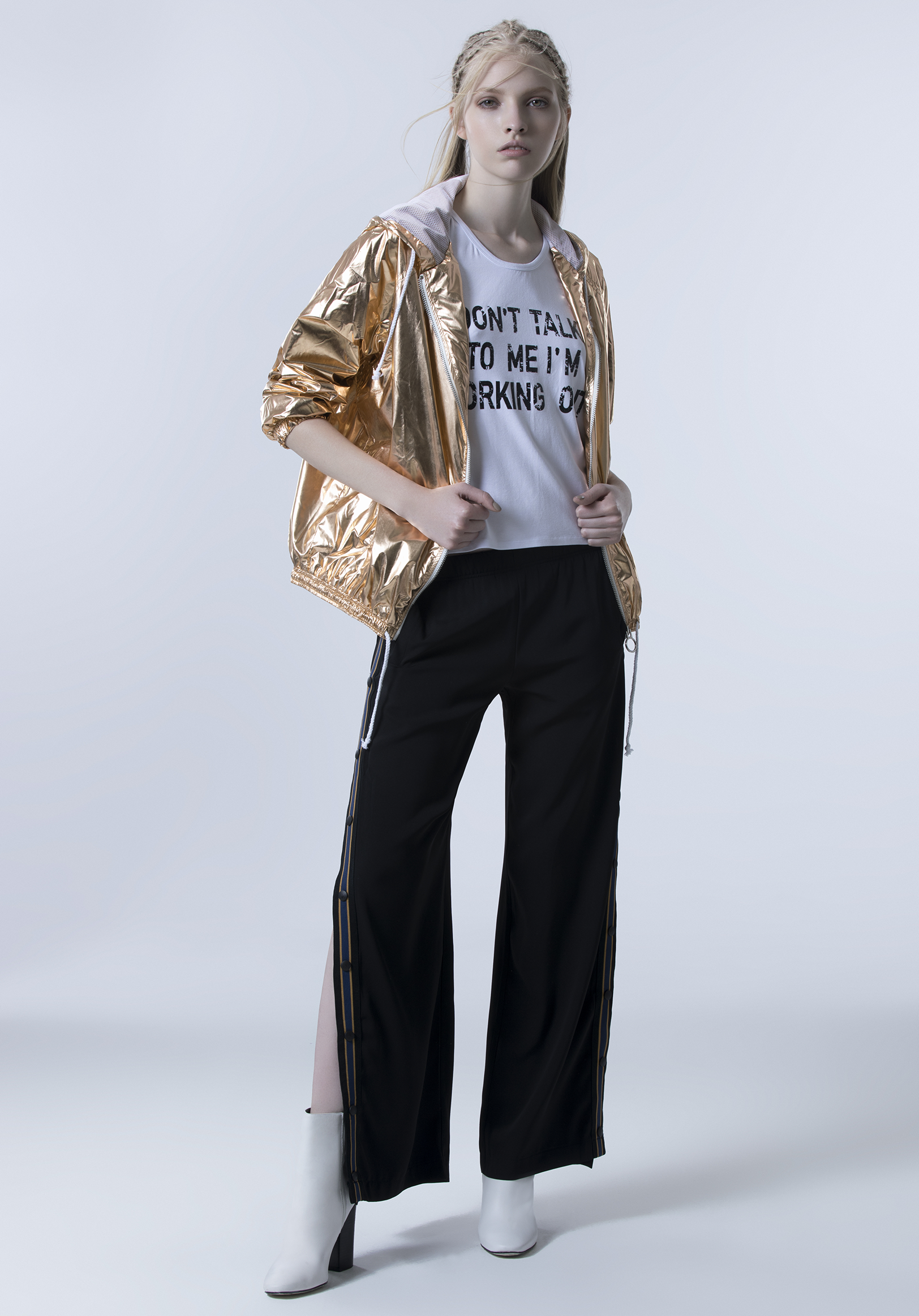 A female mannequin dressed in a white T-shirt with black text, a metallic gold jacket, black track pants with side zipper details, and white heeled boots, standing against a plain light gray background.
