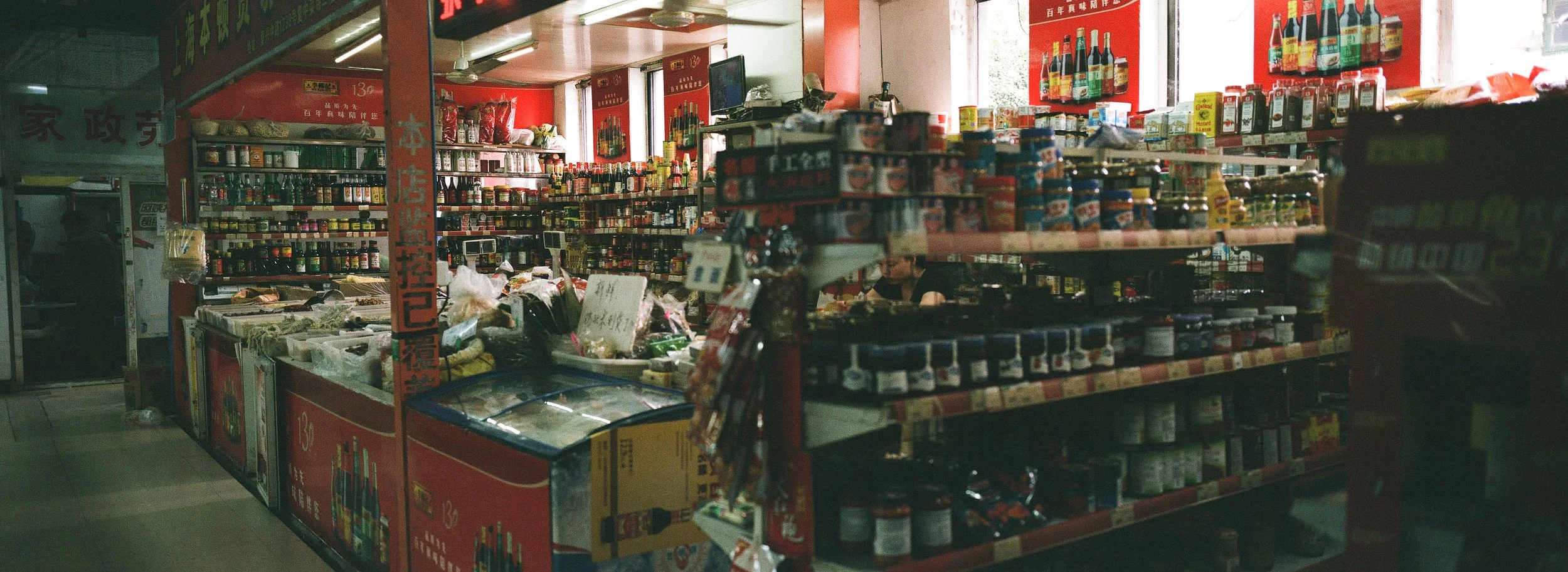 Interior of a small Asian grocery store with shelves stocked with sauces, spices, and canned goods, and a counter with refrigerated items.