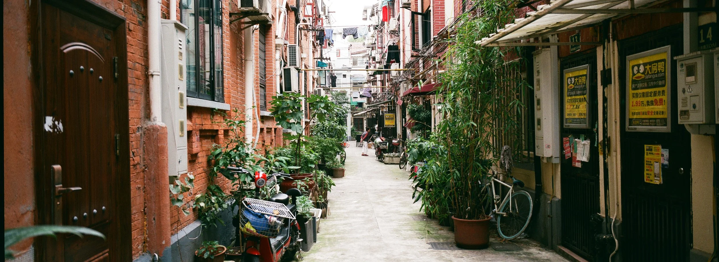 Narrow alleyway with brick buildings on both sides, plants, bicycles, and laundry hanging overhead.