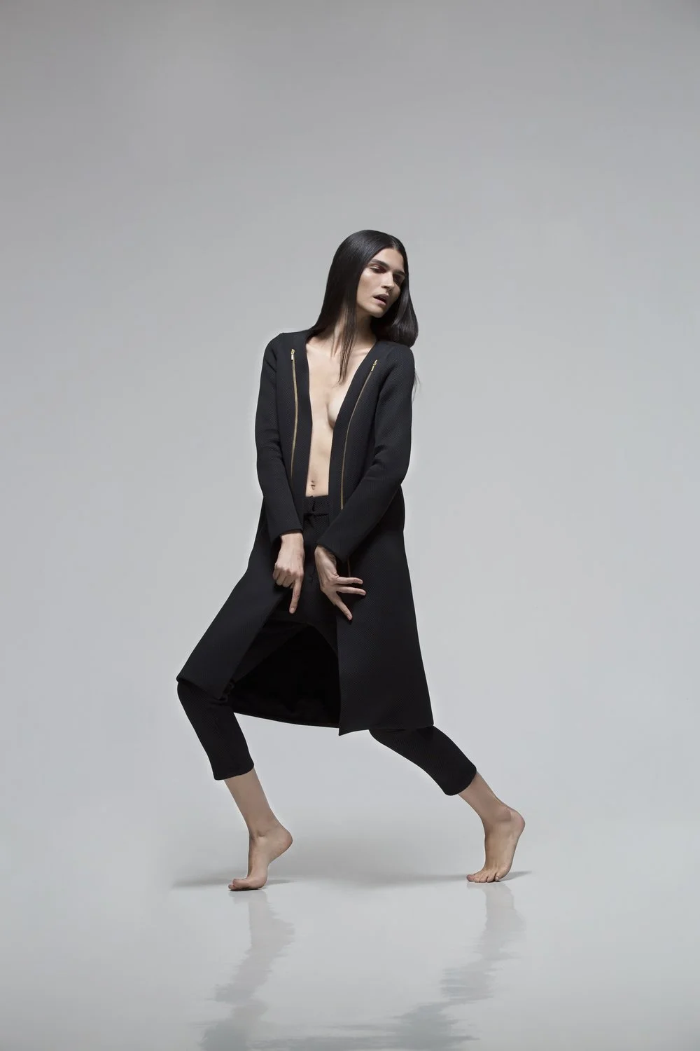 A woman with long dark hair posing barefoot in a black jacket and pants against a plain light gray background.