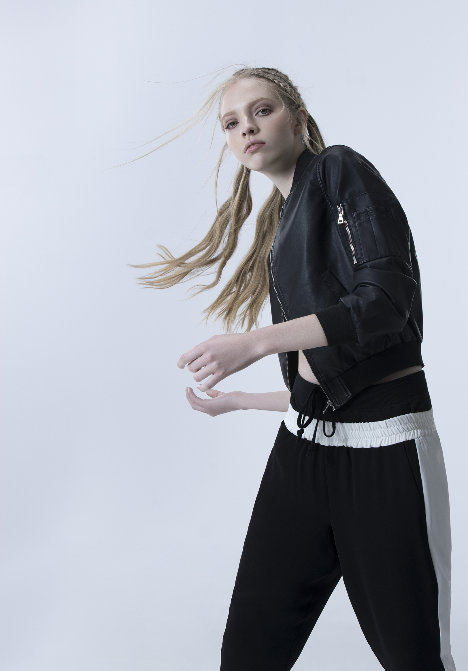 A young woman with long blonde hair in braids wearing a black leather jacket and black and white pants, standing against a plain light background.