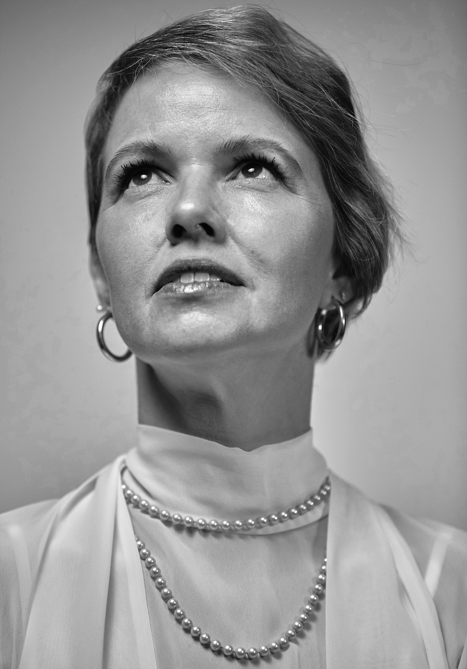 Black and white close-up portrait of a woman with short hair, wearing pearl jewelry, looking upward.