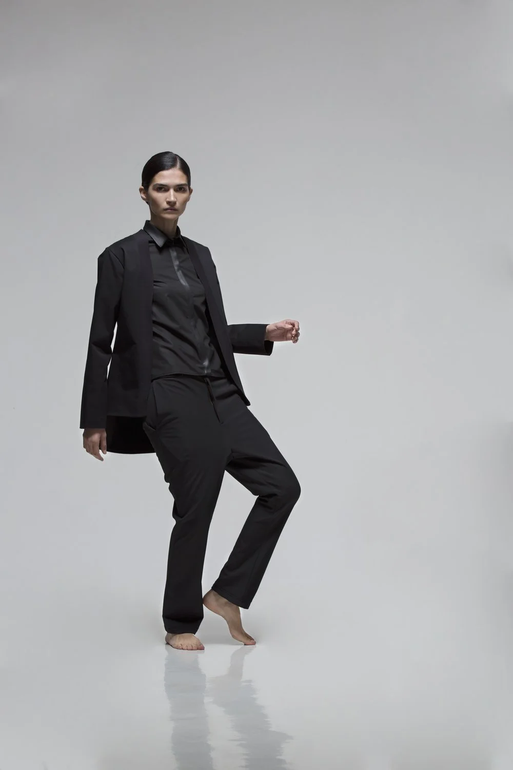 A woman dressed in black pants, black shirt, and blazer standing barefoot against a plain gray background.