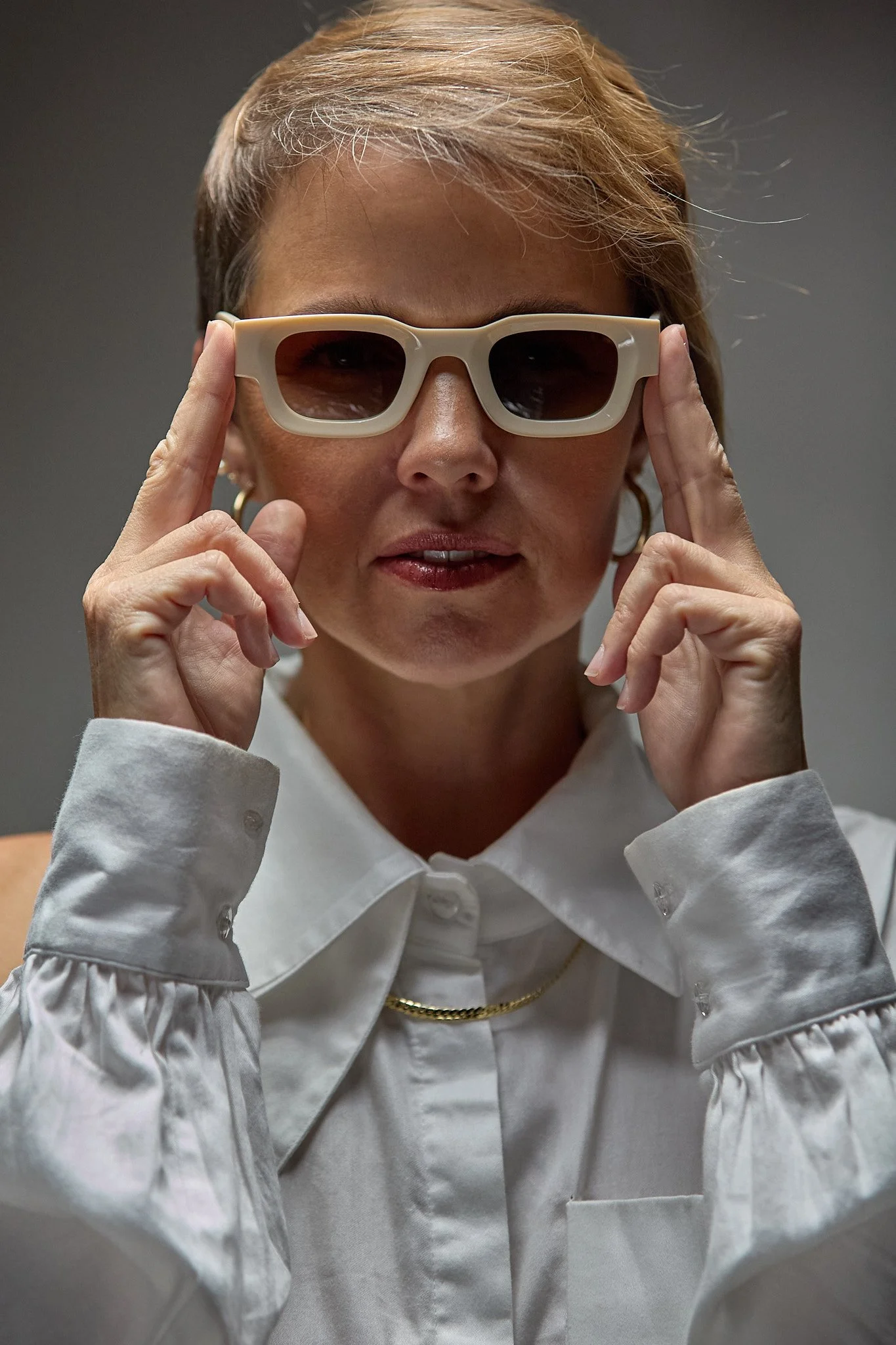 A woman wearing large white sunglasses and a white long-sleeved blouse with puffed sleeves, holding the sunglasses with both hands, with a gray background.