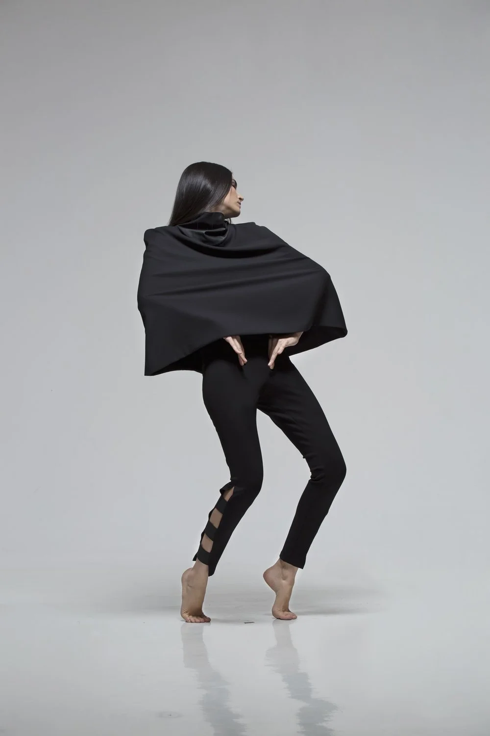 A woman with long dark hair wearing a black outfit and cape, posing barefoot against a light gray background.