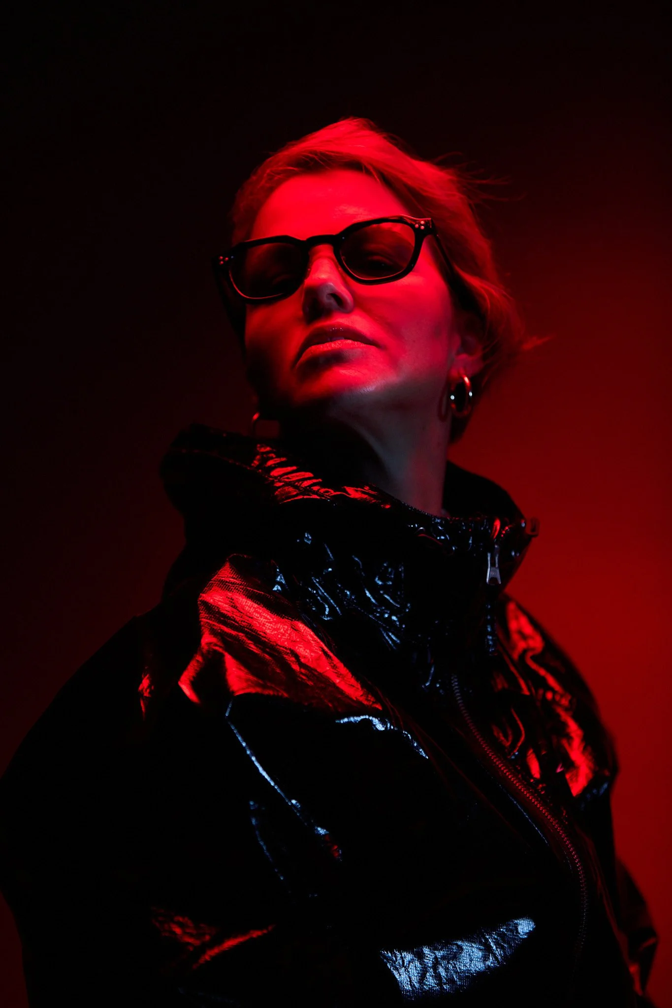 A woman with short blonde hair wearing sunglasses and a shiny black jacket, illuminated by red lighting.