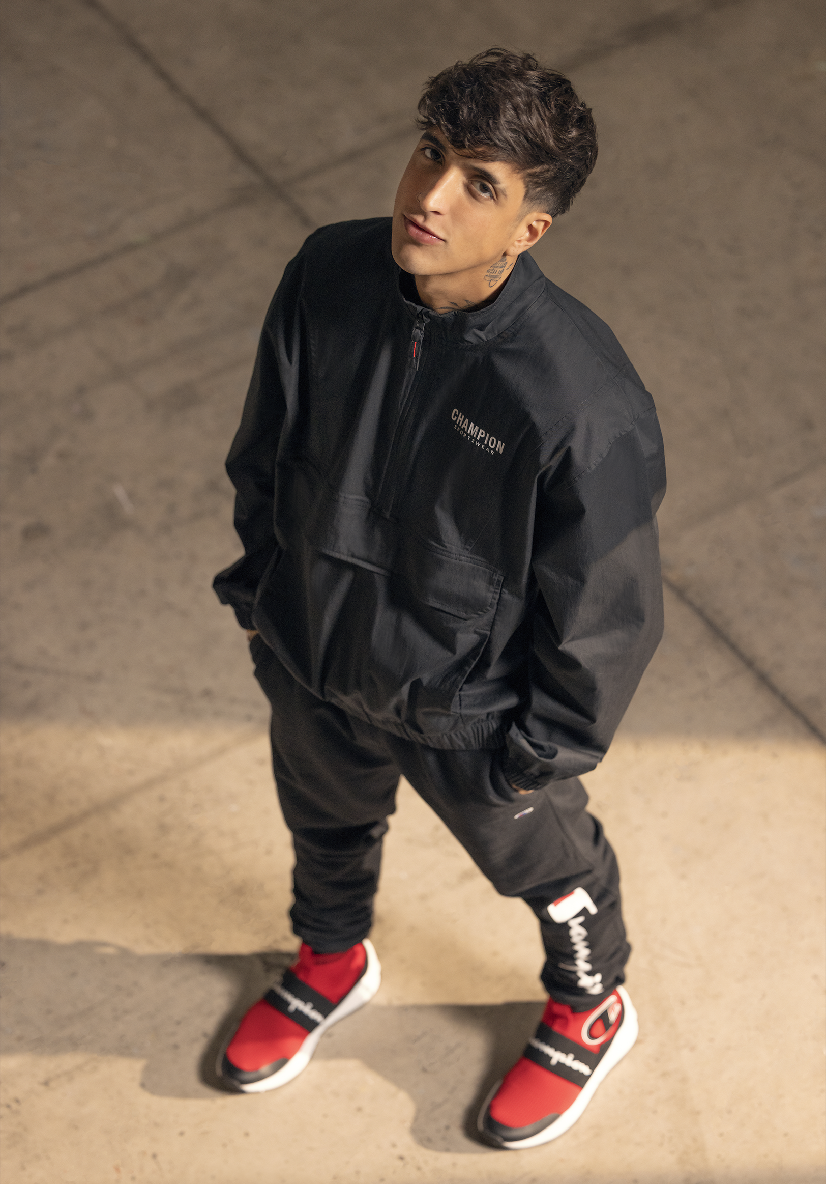 Young man with dark curly hair and tattoos on his neck, wearing a black Champion jacket, black pants, and red and black sneakers, standing on a concrete floor and looking up at the camera.