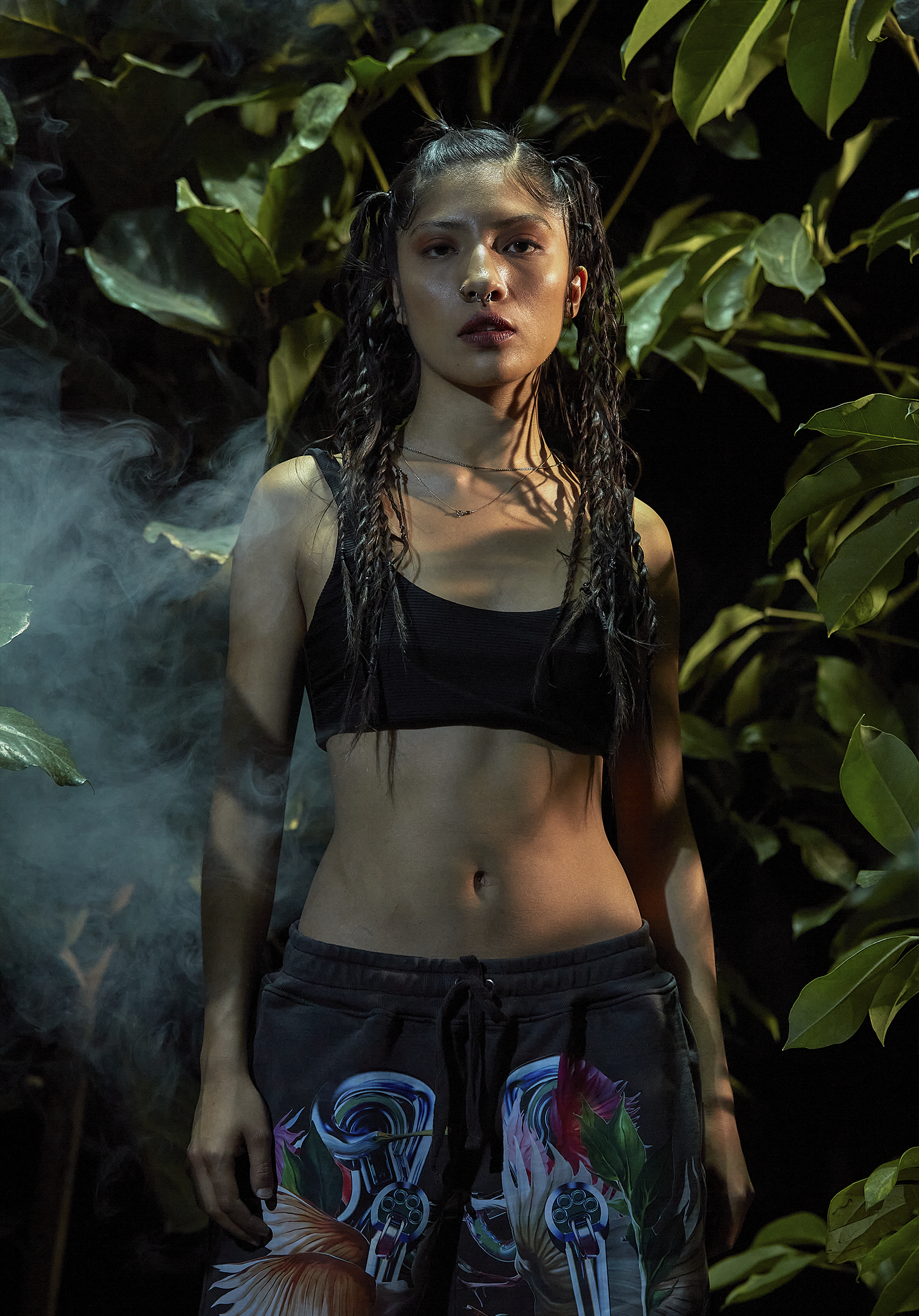 A young woman with long dark curly hair styled in pigtails standing in front of large green leaves, wearing a black crop top and colorful shorts with a design featuring revolvers and swirling patterns, with a neutral expression on her face.