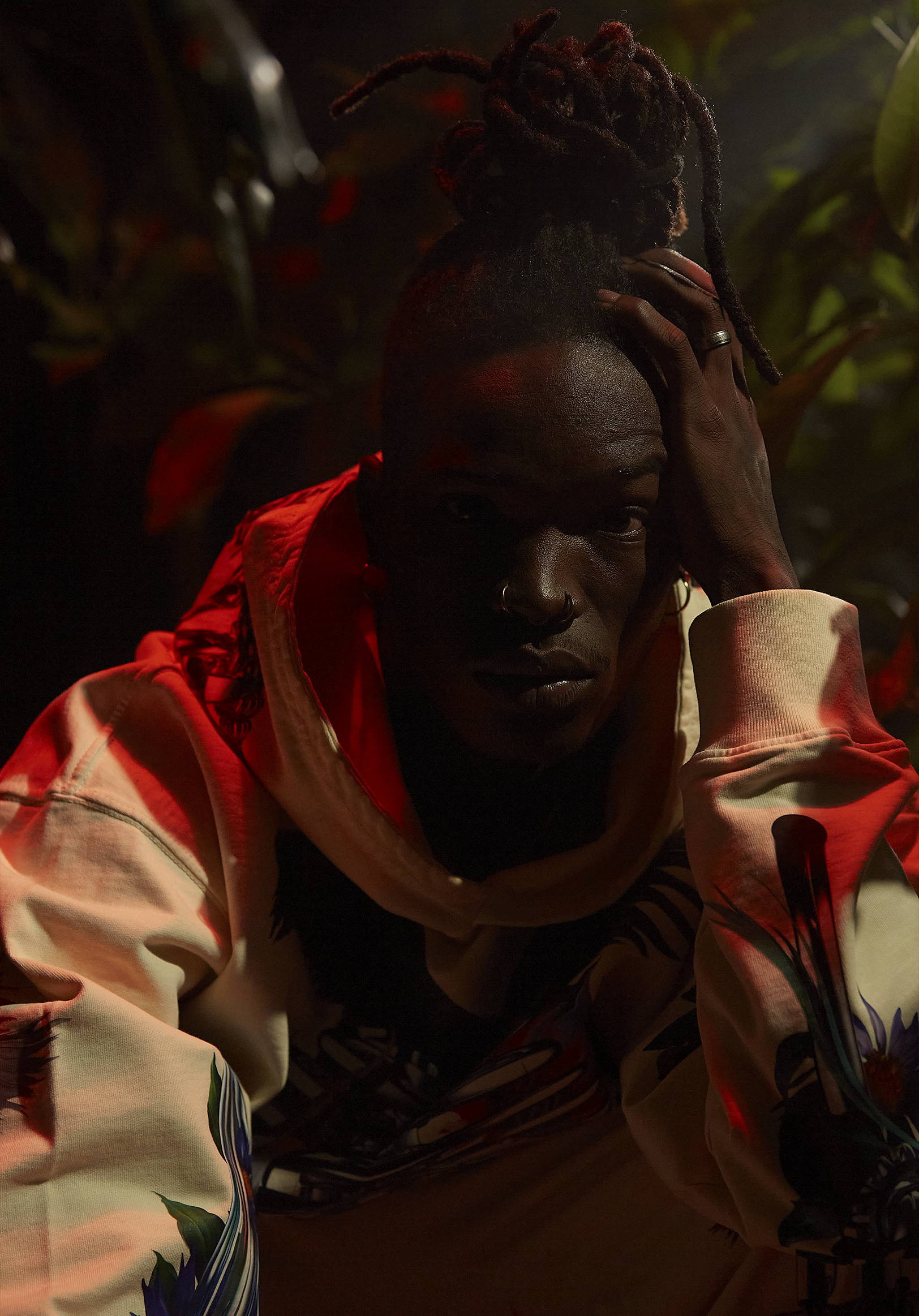 A person with dreadlocks and dark skin, wearing a colorful jacket, resting their head on their hand in a dimly lit environment with green foliage in the background.