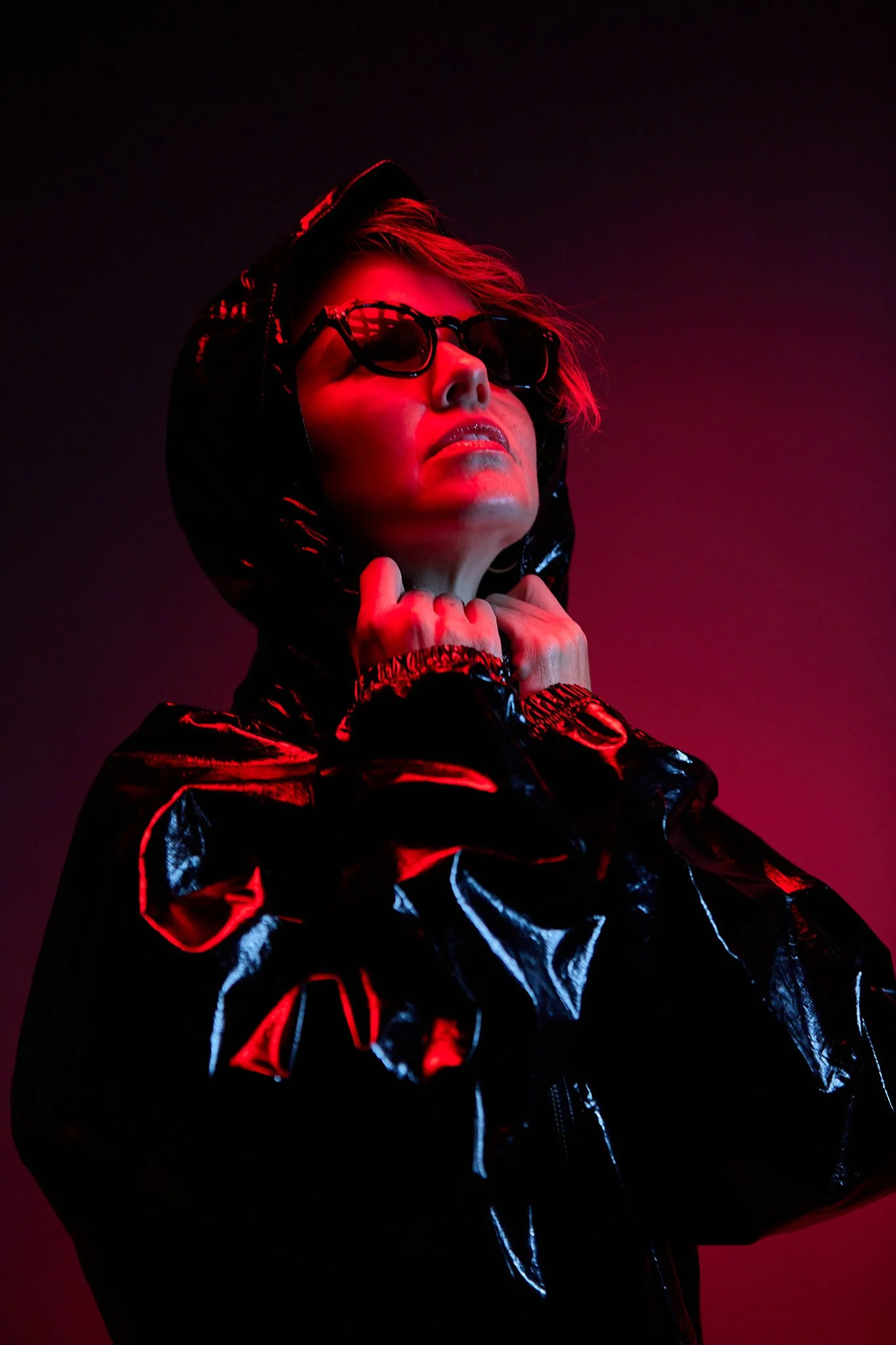 A person wearing black sunglasses and a shiny black jacket with red and blue highlights, with a dark background and red lighting on their face and clothing