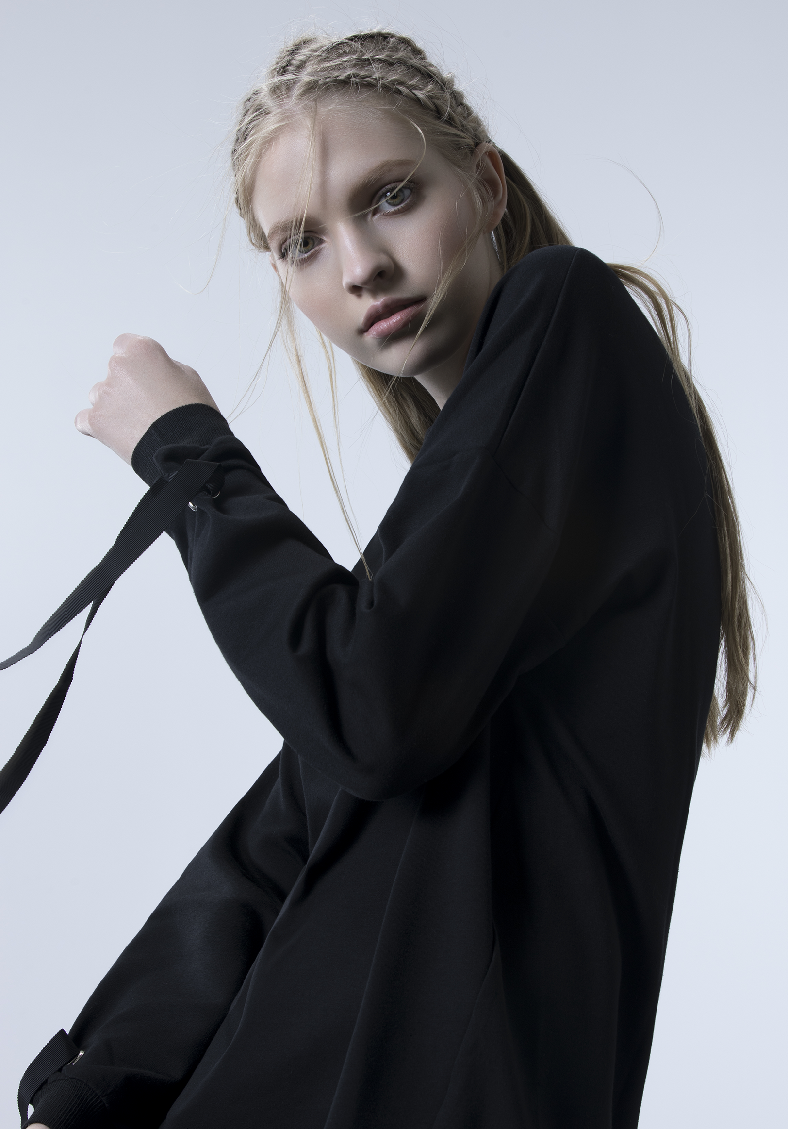 Portrait of a young woman with blonde hair styled in braids, wearing a black hoodie, posing against a plain gray background.