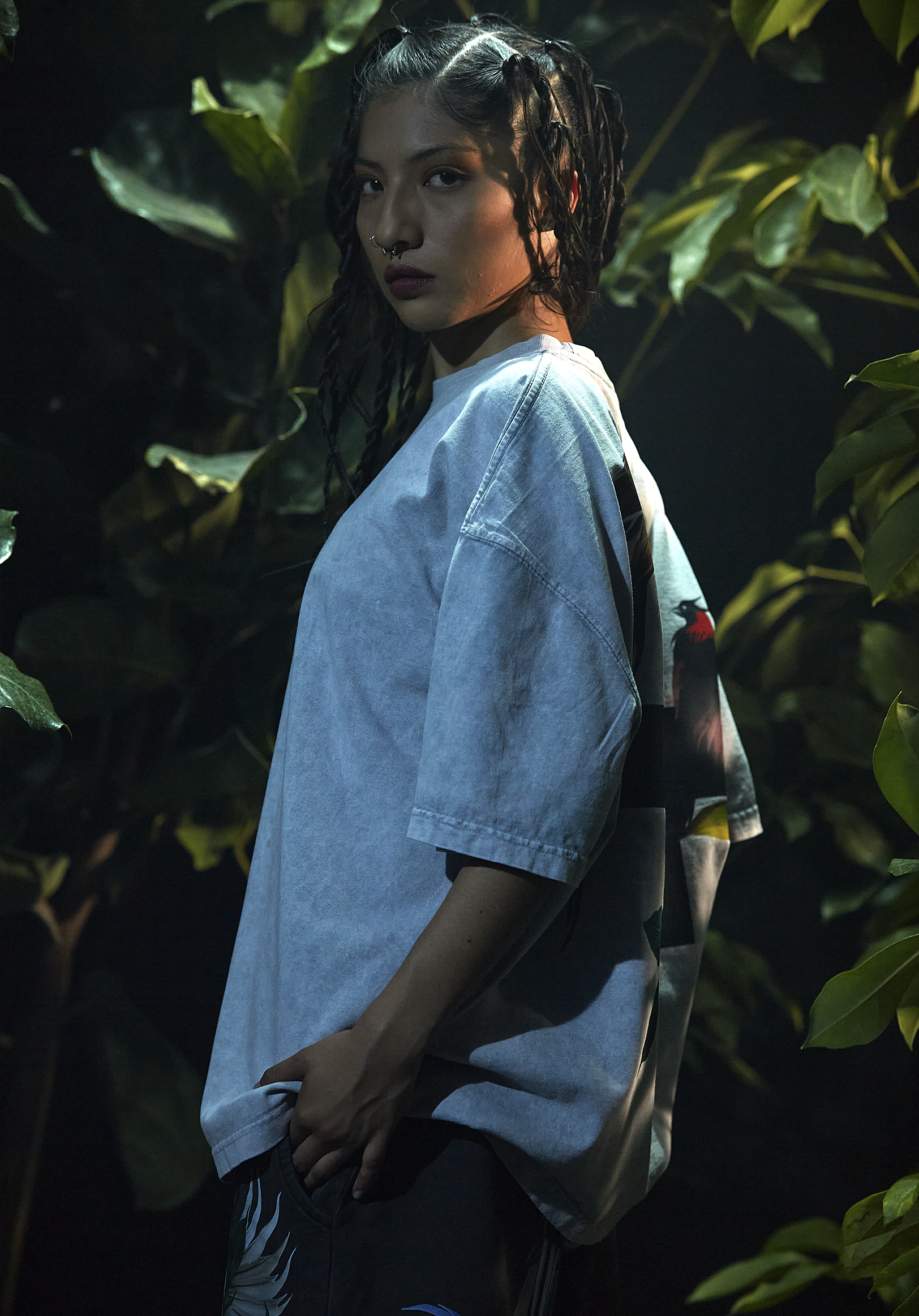 A person with curly hair and a septum piercing standing among dark green leaves at night, wearing a light-colored oversized shirt.