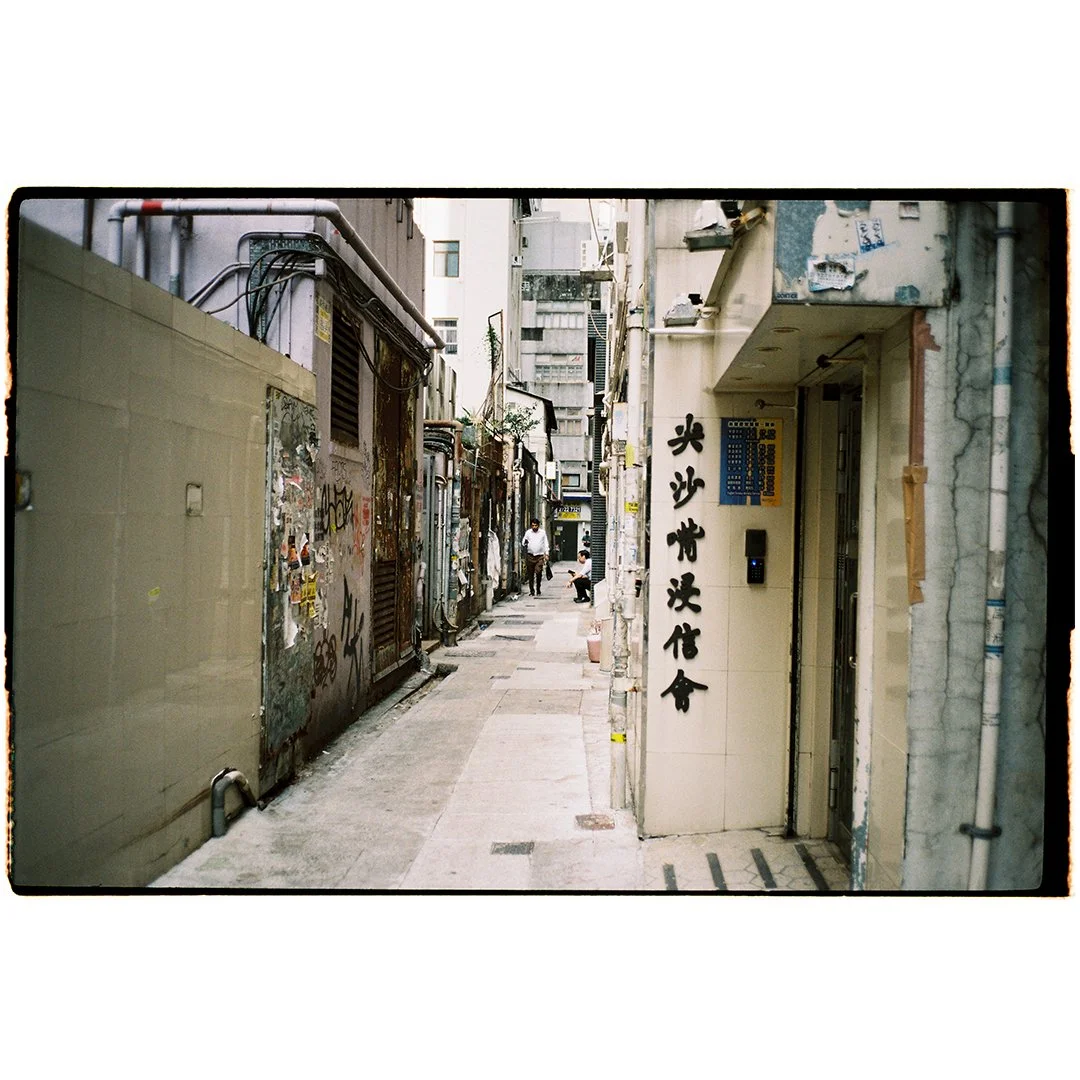 Narrow urban alleyway with graffiti-covered walls on the left and a building with Chinese characters on the right. Two people are sitting and standing further down the alley.