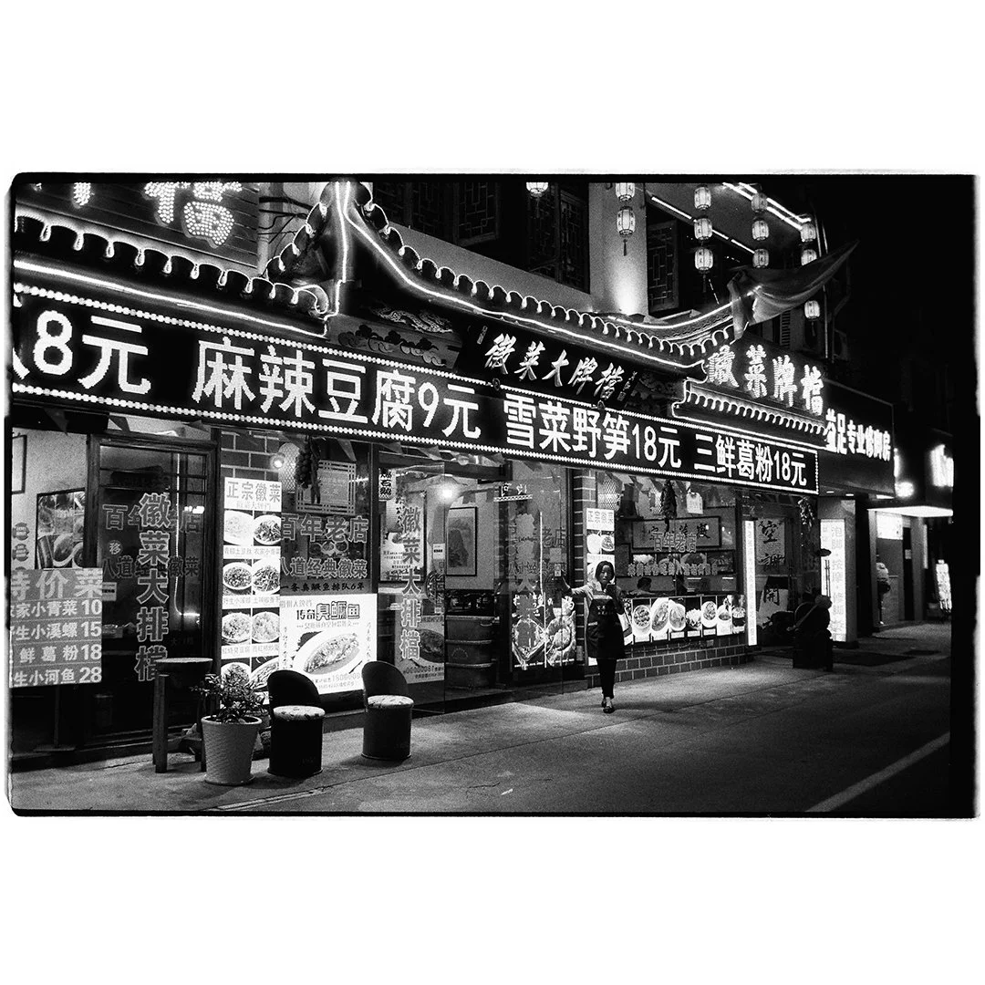 Night scene of a Chinese street food restaurant with illuminated Chinese signs and menus, featuring various dishes and prices visible in Chinese characters.