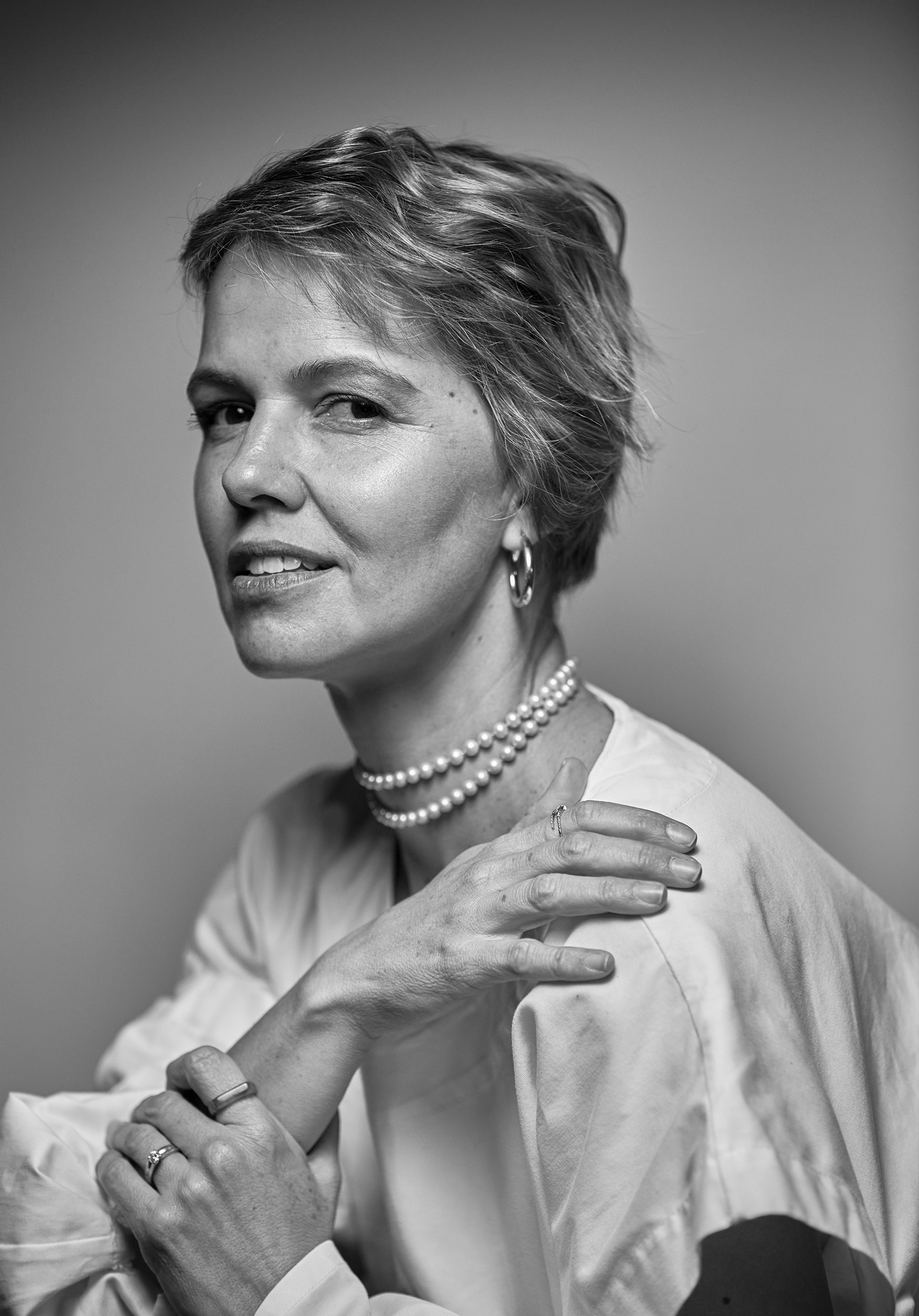 Black and white portrait of a woman with short hair, wearing a pearl necklace and earrings, with her hand resting on her shoulder, looking slightly to the side and winking.