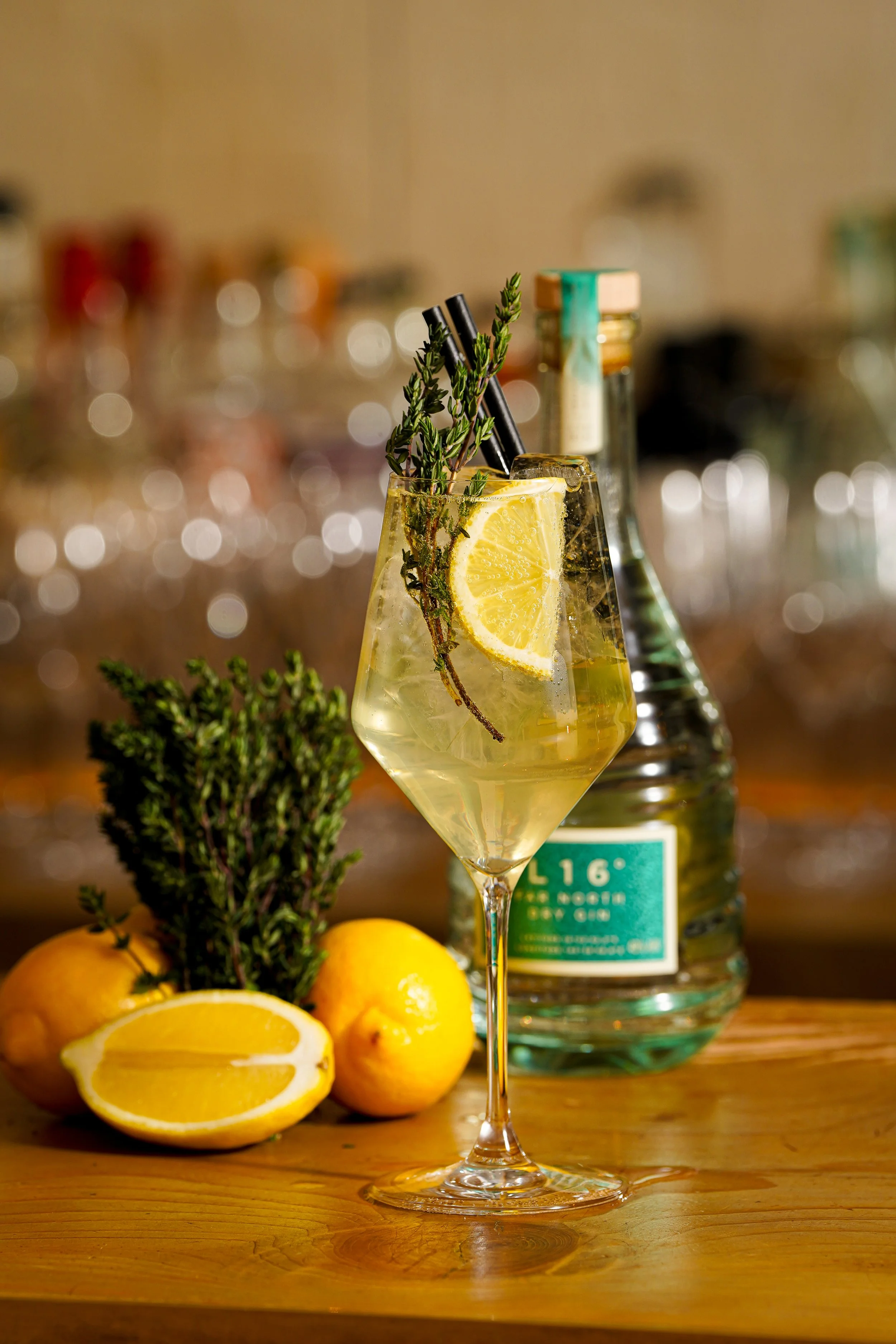 A glass of lemon and herb cocktail with lemon slices and sprigs of herbs, set on a wooden surface with fresh lemons, herbs, and a bottle in the background, at a restaurant in Avalon.